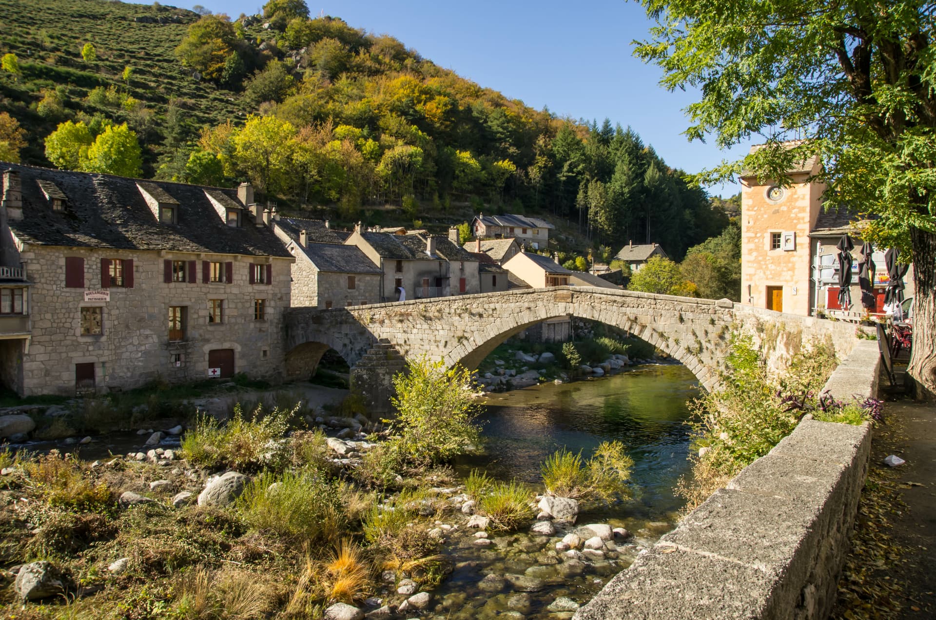 Stone bridge over river in Pont-de-Montvert village with autumn foliage on hillside.