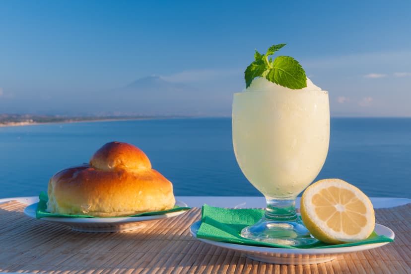 Granita drink with mint, brioche bun, and lemon slice with sea and distant mountain background