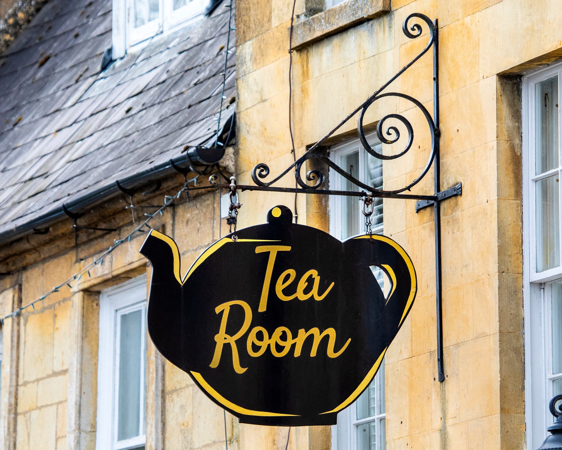 Black teapot-shaped "Tea Room" sign hanging on a yellow stone building in Moreton-in-Marsh.