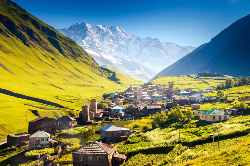 Alpine village with stone houses and watchtowers in a green mountain valley beneath snow-capped peaks