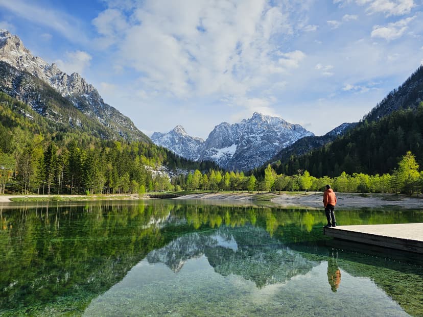 Person standing on wooden dock at alpine lake with snowy mountains reflection