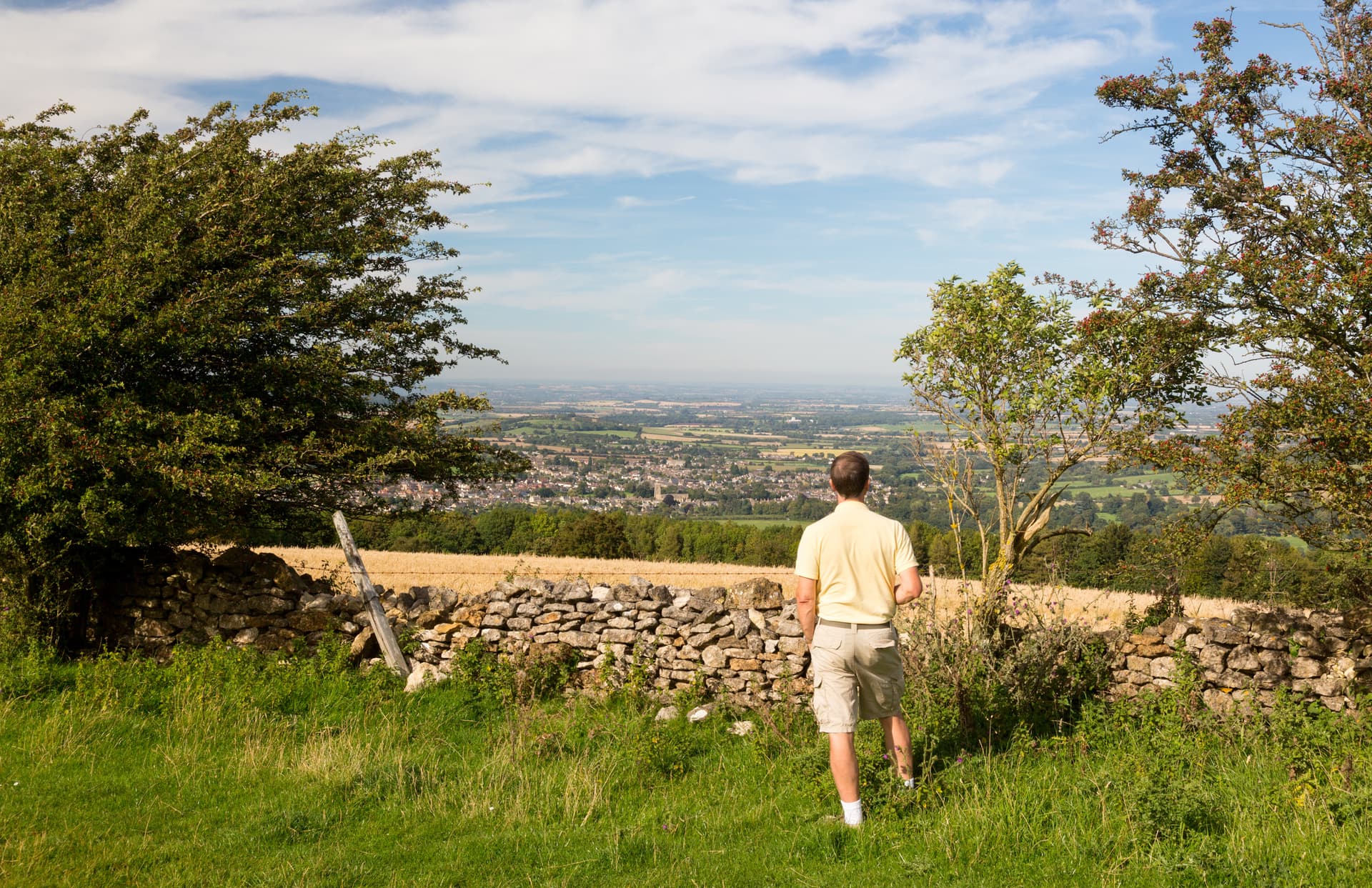 Man viewing Winchcombe valley from hillside with stone wall and trees