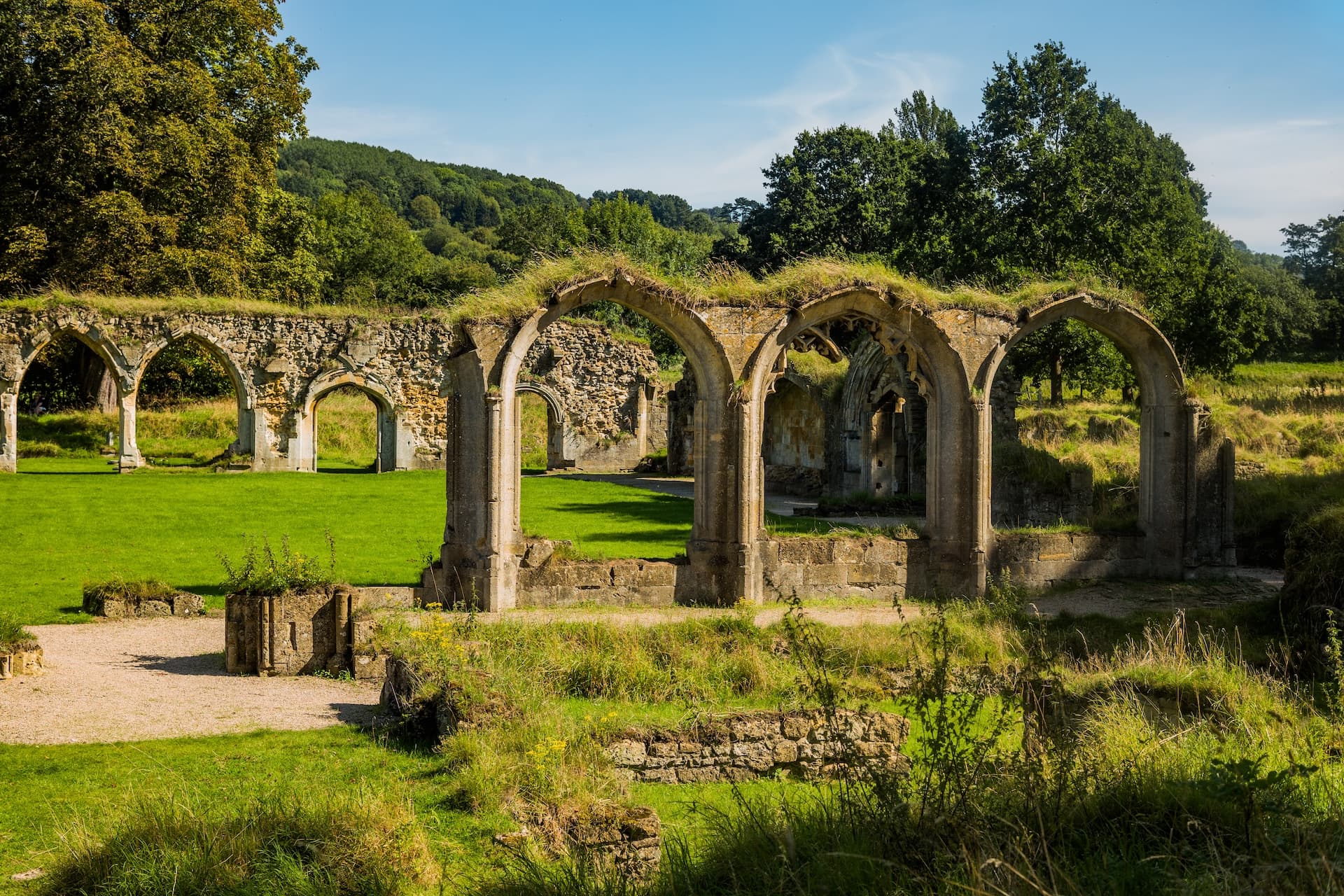 Ruins of Hailes Abbey with Gothic arches, green grass, and wooded hills under a blue sky.