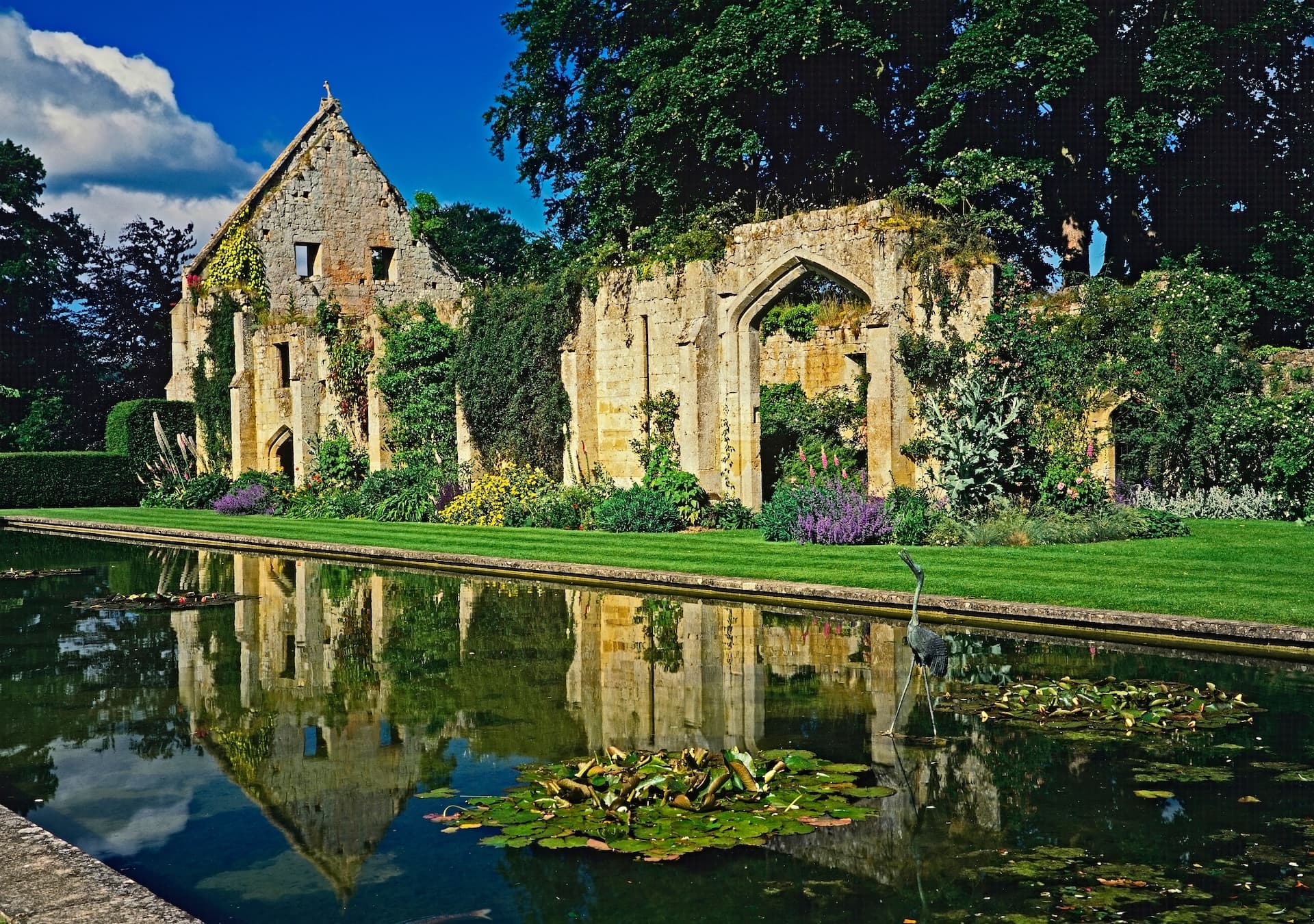Sudeley Castle ruins reflected in a long pond with a heron wading near lily pads.
