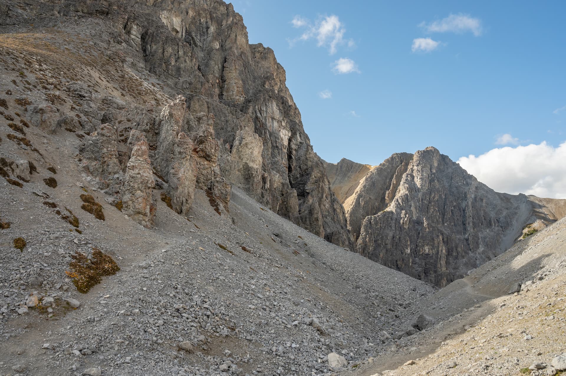 Rocky mountain pass with scree slopes and a narrow trail under a blue sky.