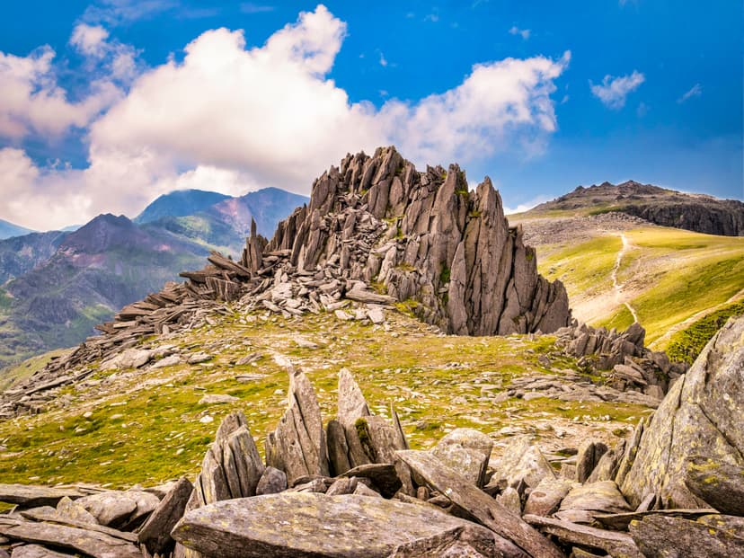 Rugged rocky mountain peak with jagged stone formations and grassy slopes under a blue sky with white clouds