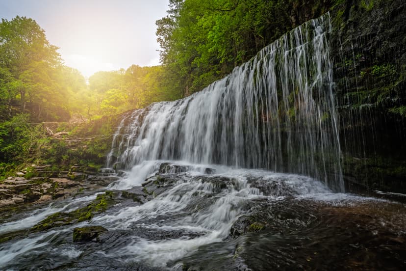 Waterfall cascading over a rocky cliff with lush green trees and sunlight