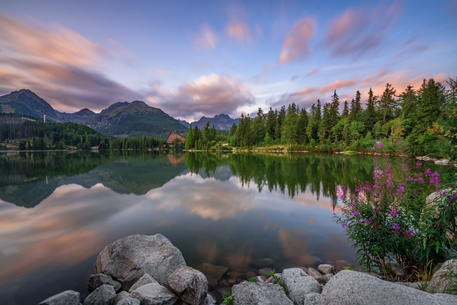 Alpine lake Štrbské Pleso reflecting mountains and colorful sunset clouds, with purple flowers.