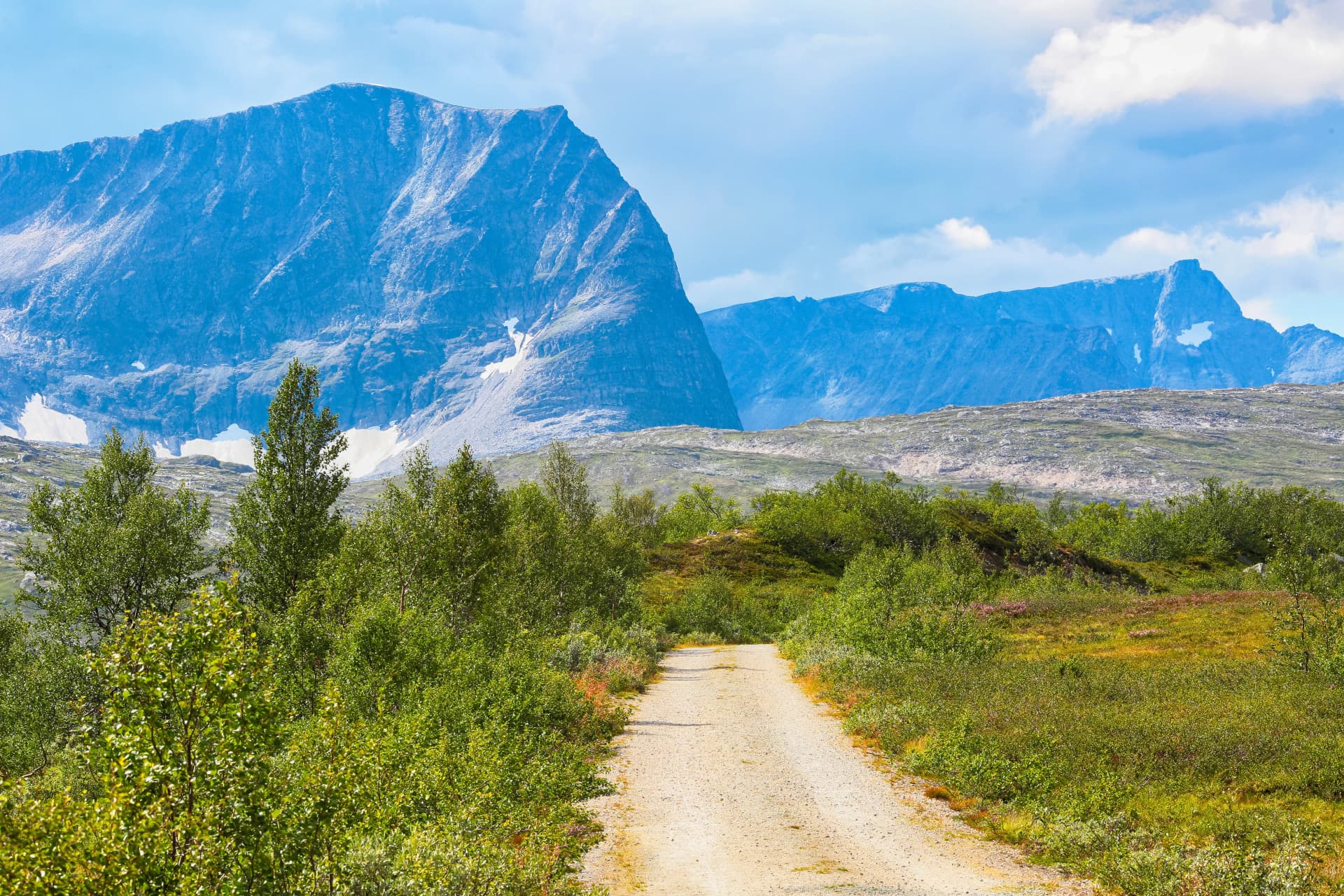 Gravel road through green scrub leading toward massive blue mountains with summer snow patches in Trollheimen, Norway.