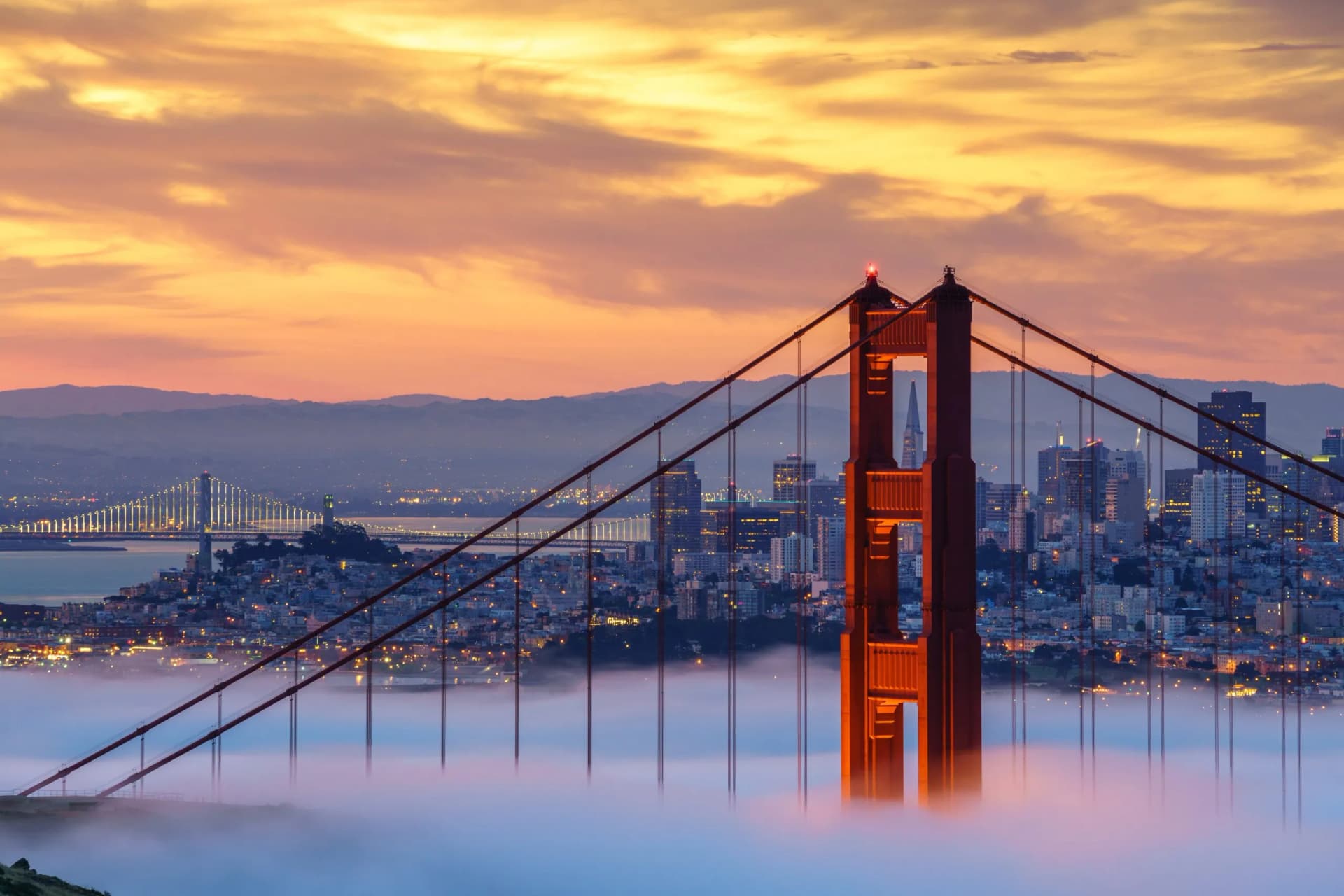 Golden Gate Bridge tower above low fog with San Francisco skyline and Bay Bridge at sunset