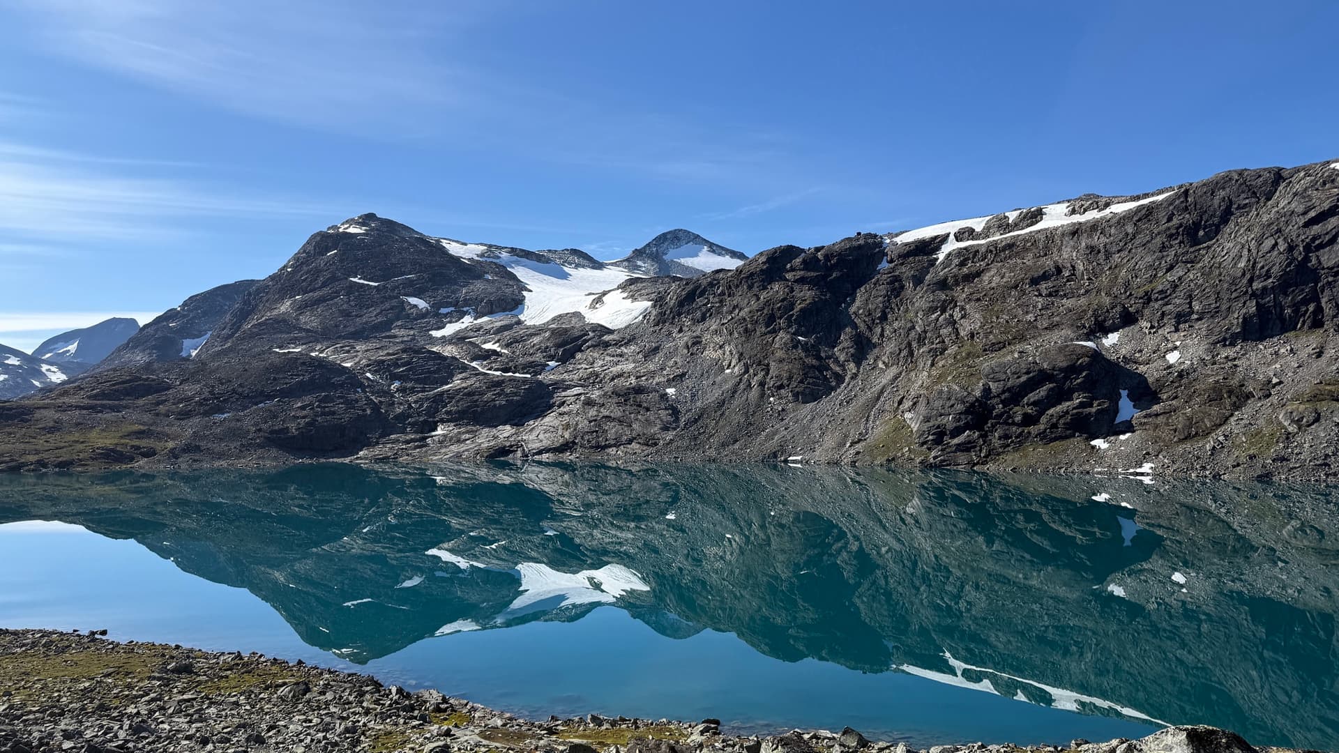 Alpine lake mirroring rugged mountain peaks with snow patches under clear blue sky