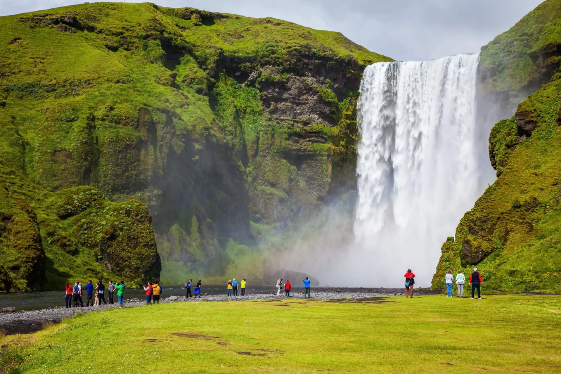 Skogafoss waterfall cascading down mossy cliffs with tourists viewing on green field.
