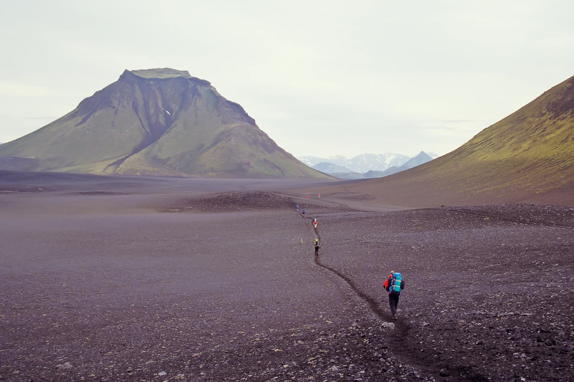 Hikers trekking across a black ash field toward green mountains on the Laugavegur trail.