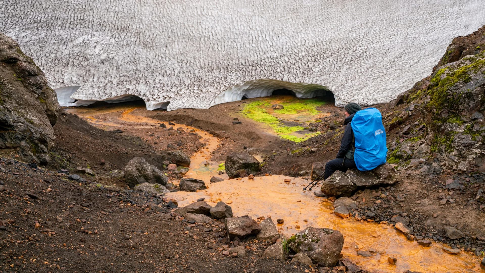 Hiker resting by orange thermal stream near melting snow cave near Kaldaklofsjokull glacier.