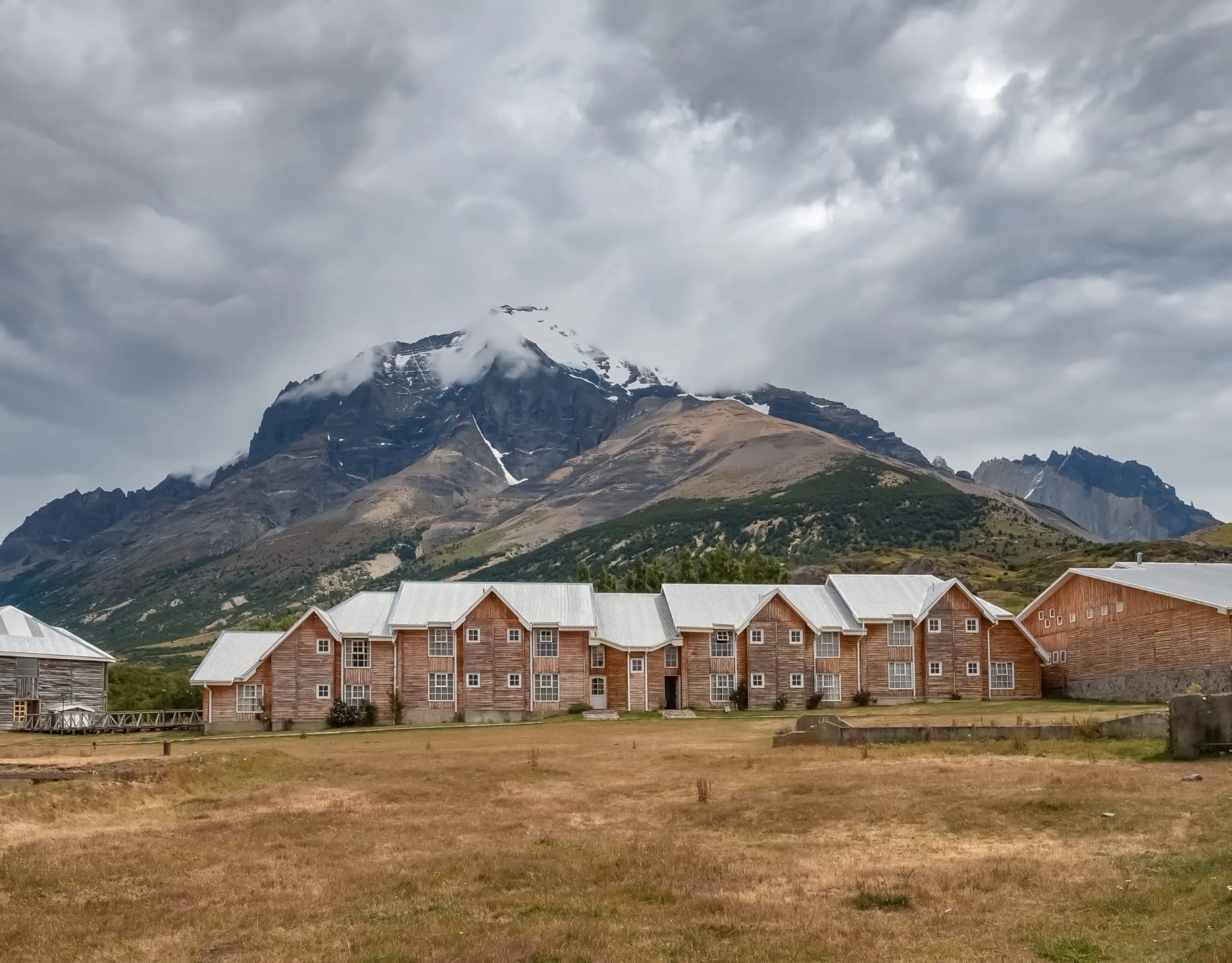 Wooden lodge buildings before dramatic snow-capped mountains under cloudy sky in Paine Massif.