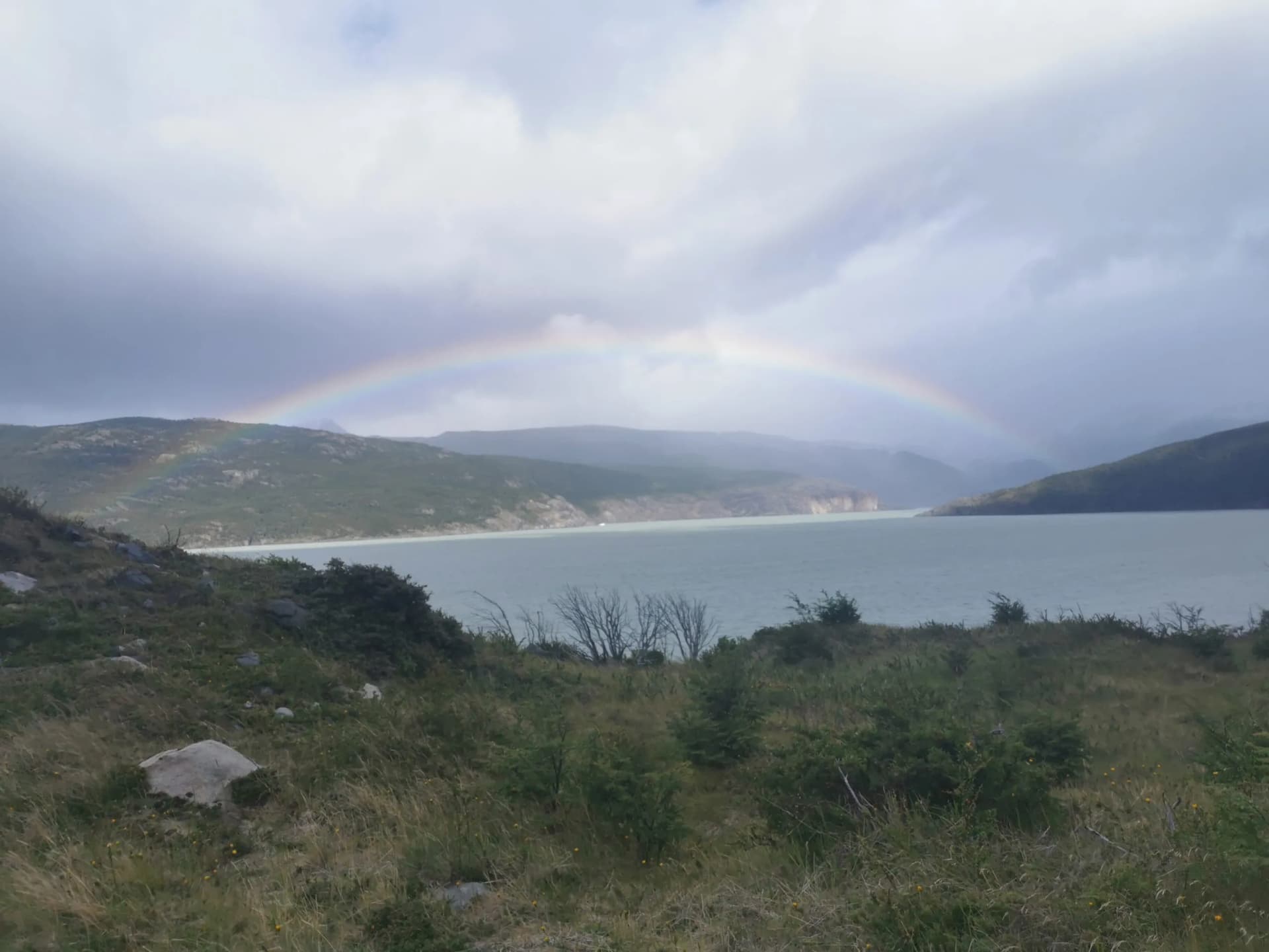 Rainbow over a lake or fjord surrounded by green, rolling hills under a cloudy sky.