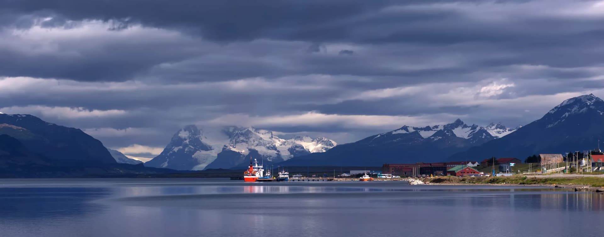 Boats docked near town with snow-capped mountains under dark clouds in Puerto Natales.