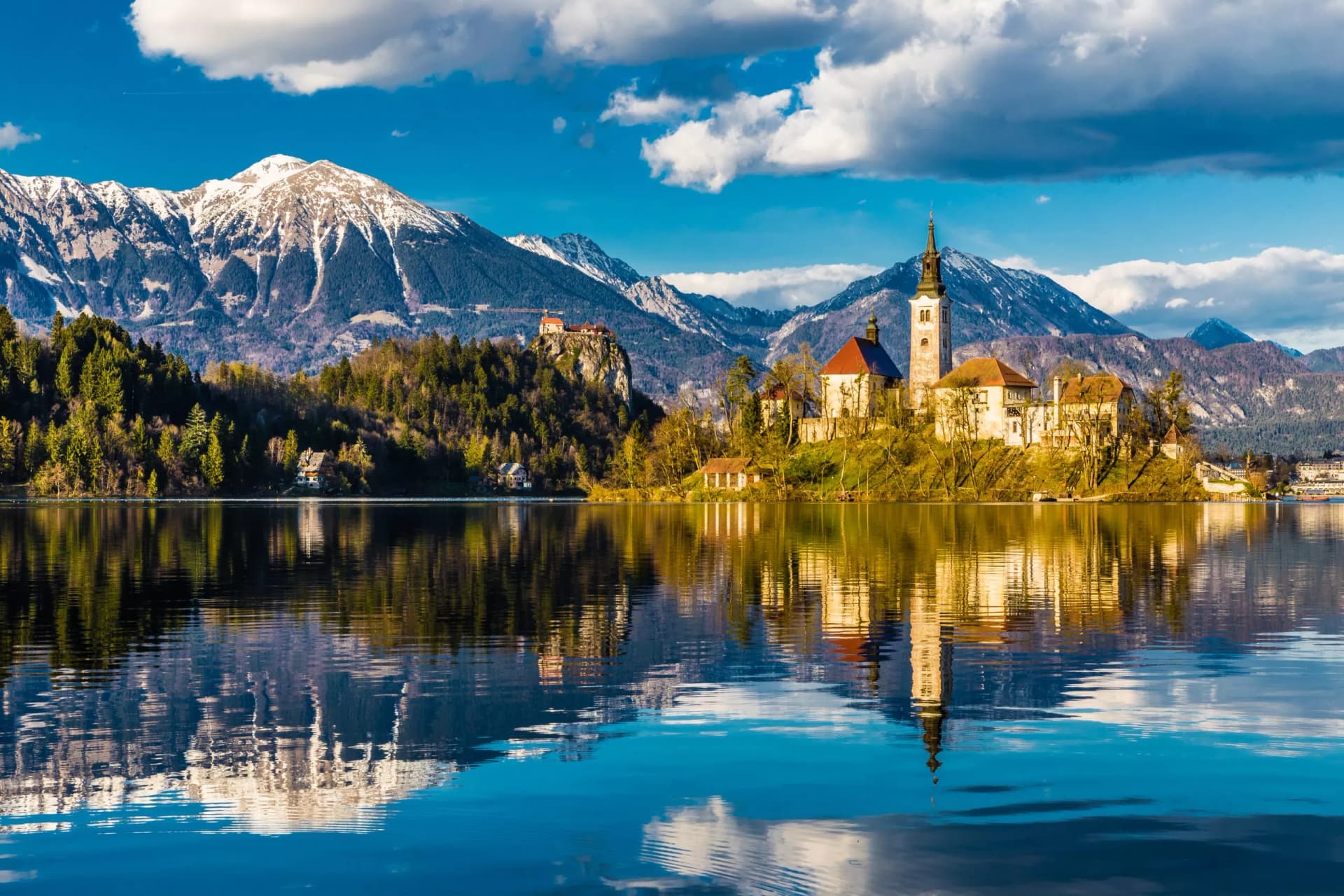Lake Bled church and castle reflected in water with snow-capped Julian Alps mountains.