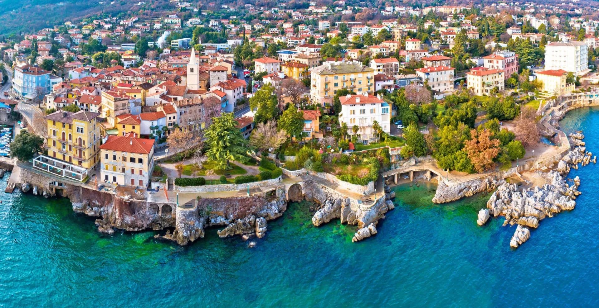Coastal town with colorful buildings above turquoise water and rocky shoreline, Lovran Lungomare.