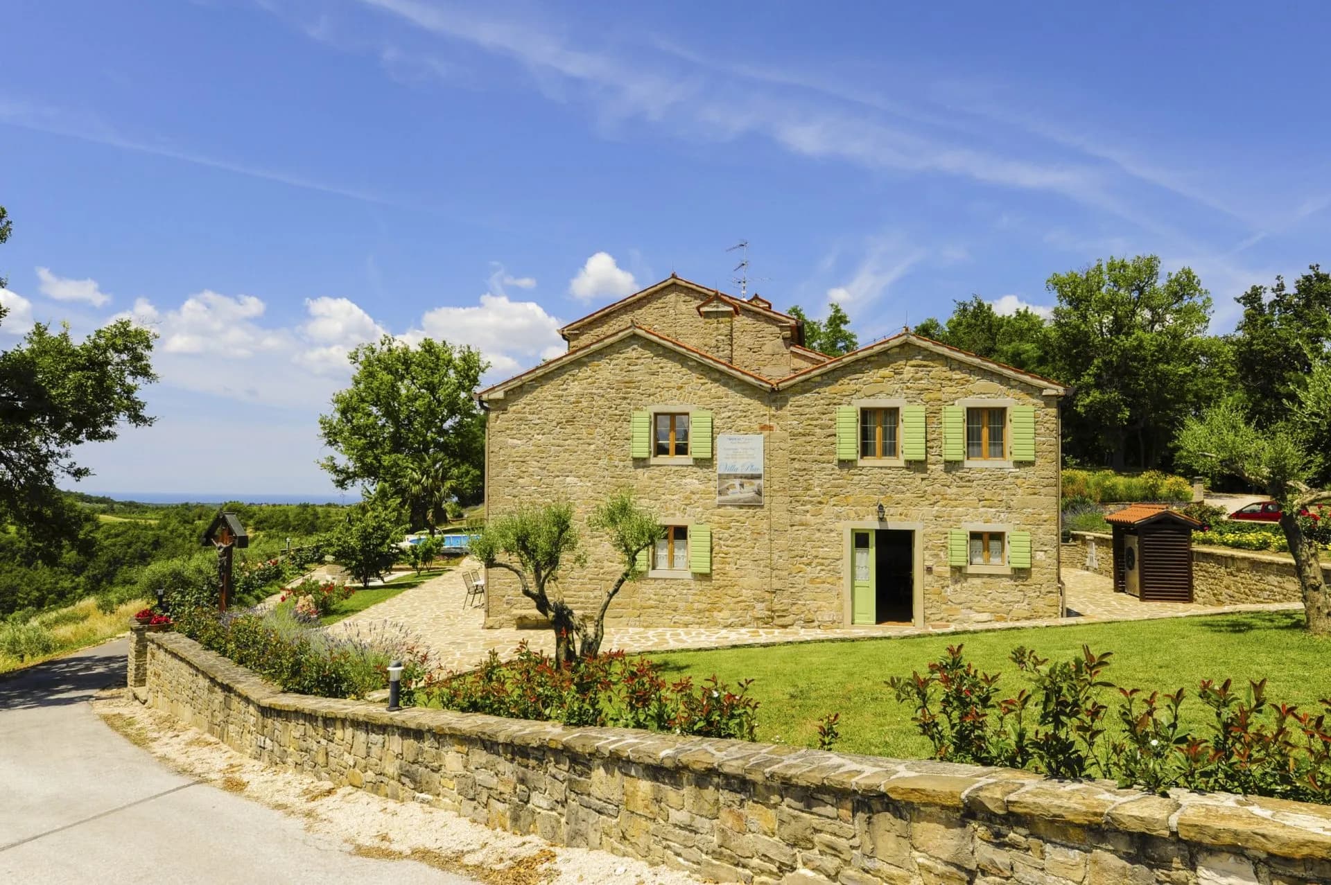 Stone house in Krasica with green shutters, lawn, and distant sea view under blue sky.