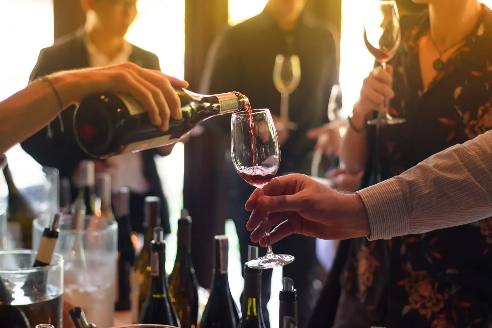 Pouring red wine during a tasting event with bottles in the foreground and people socializing.