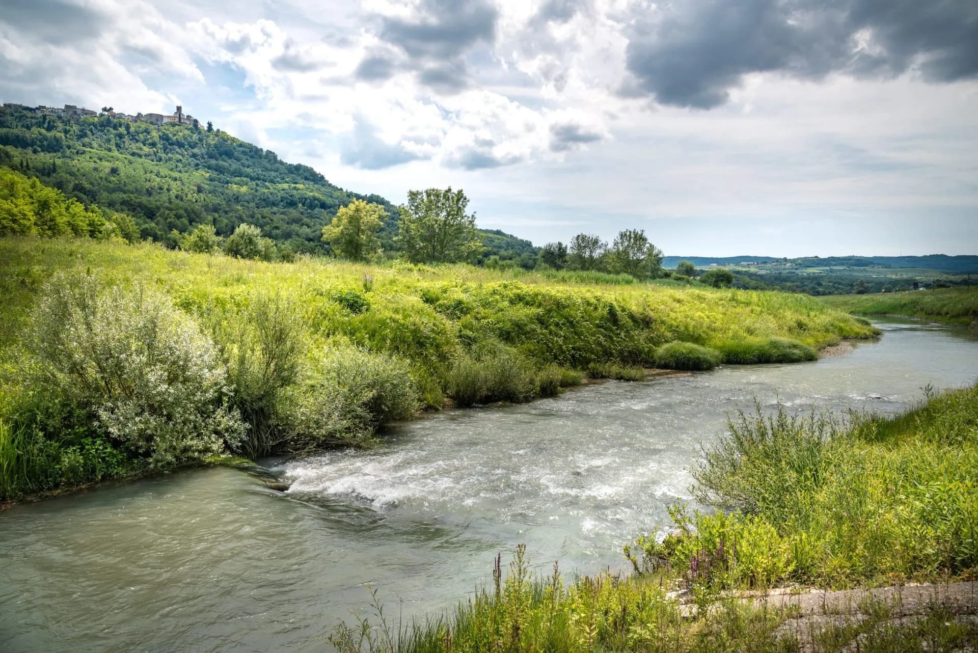 Rushing river flows past lush green banks with a hill town visible on the distant forested slope.