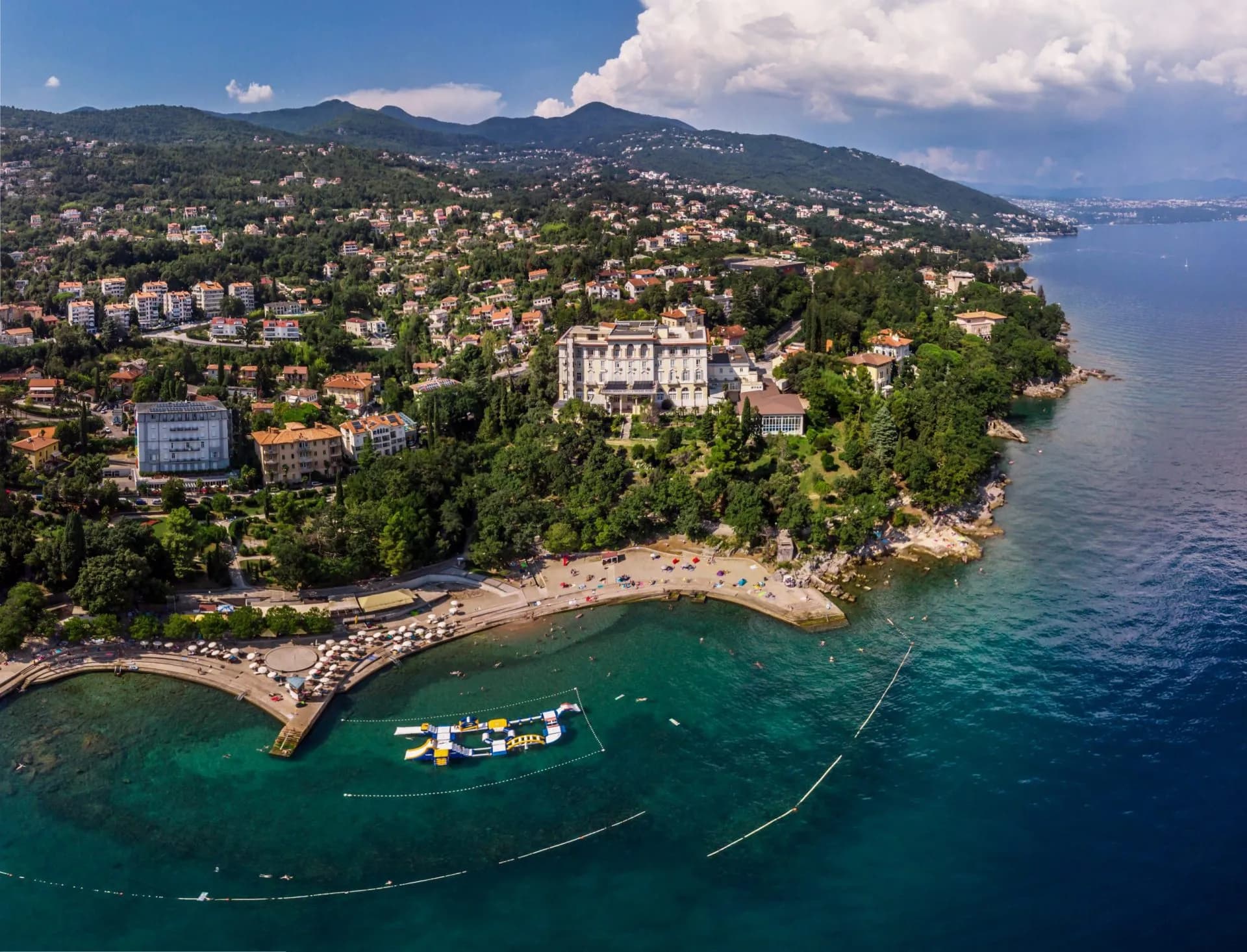 Panoramic view of Lovran coastline with beach, turquoise water, and buildings on forested hills.