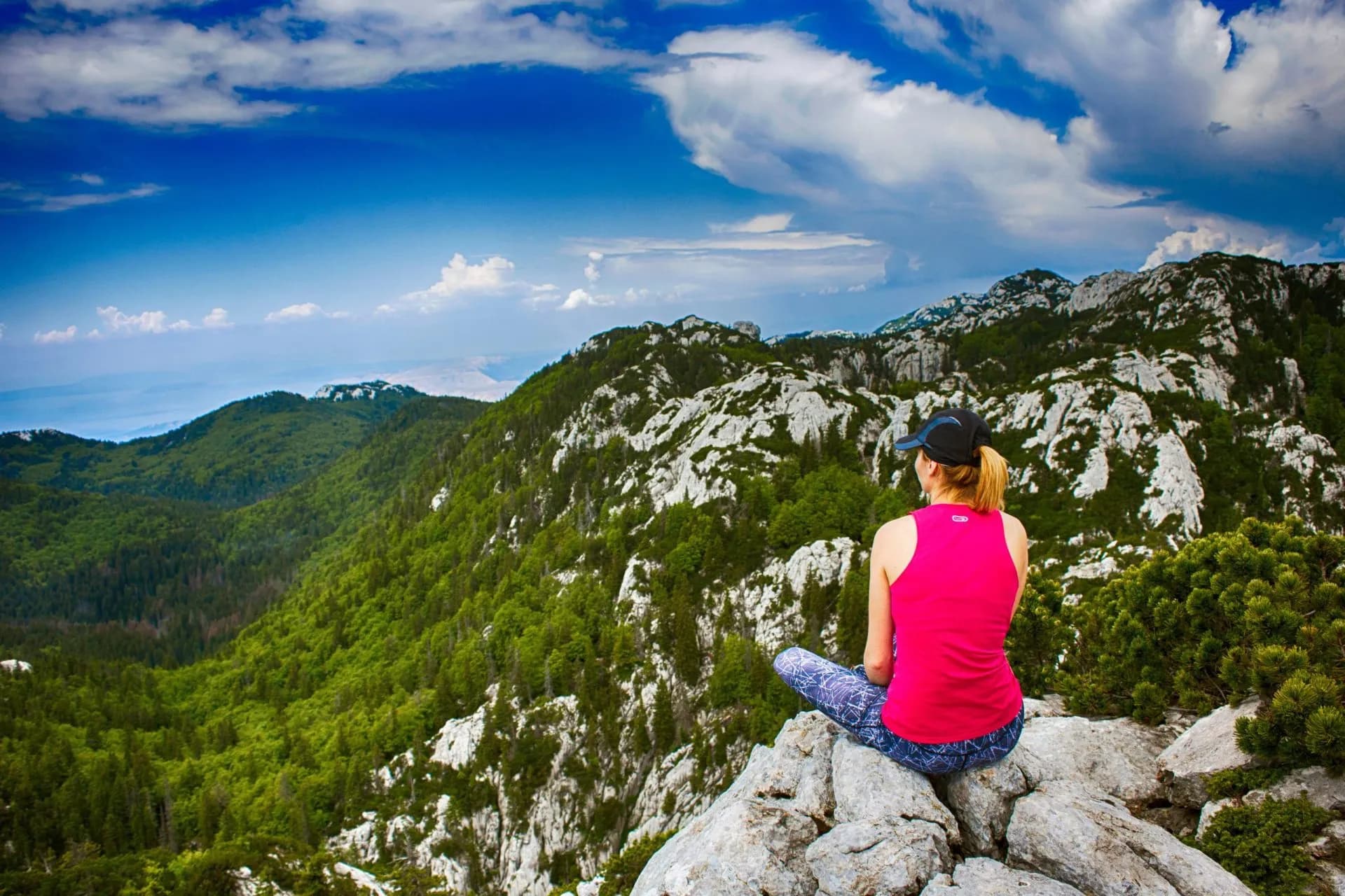 Hiker resting on rocks overlooking Velebit mountains with lush green slopes and blue sky.