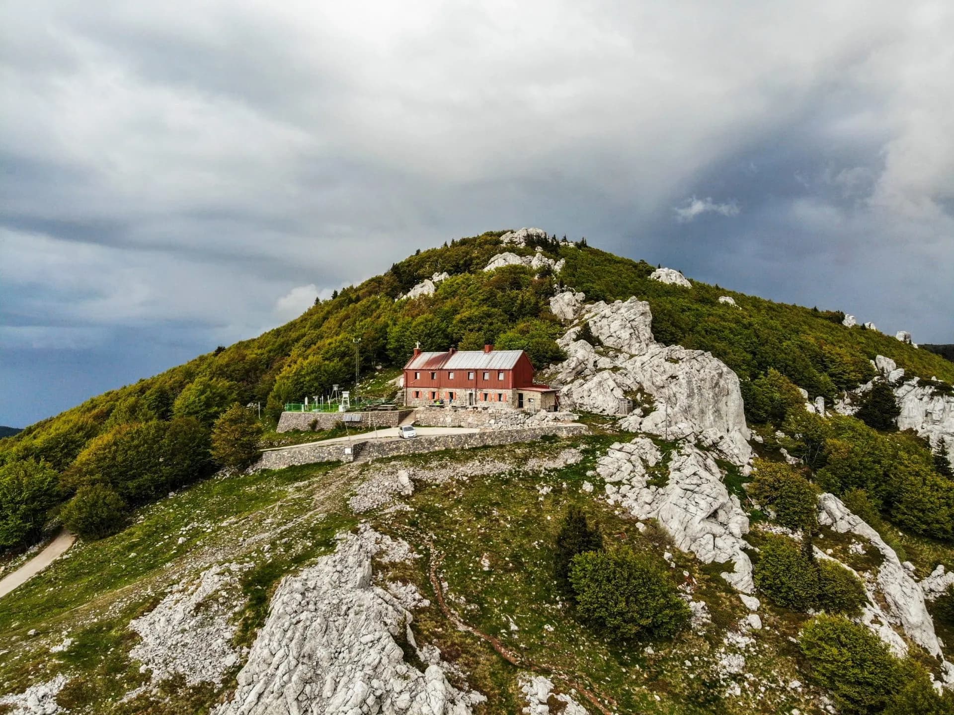 Mountain hut on rocky, green peak under stormy sky, possibly a hiking refuge.