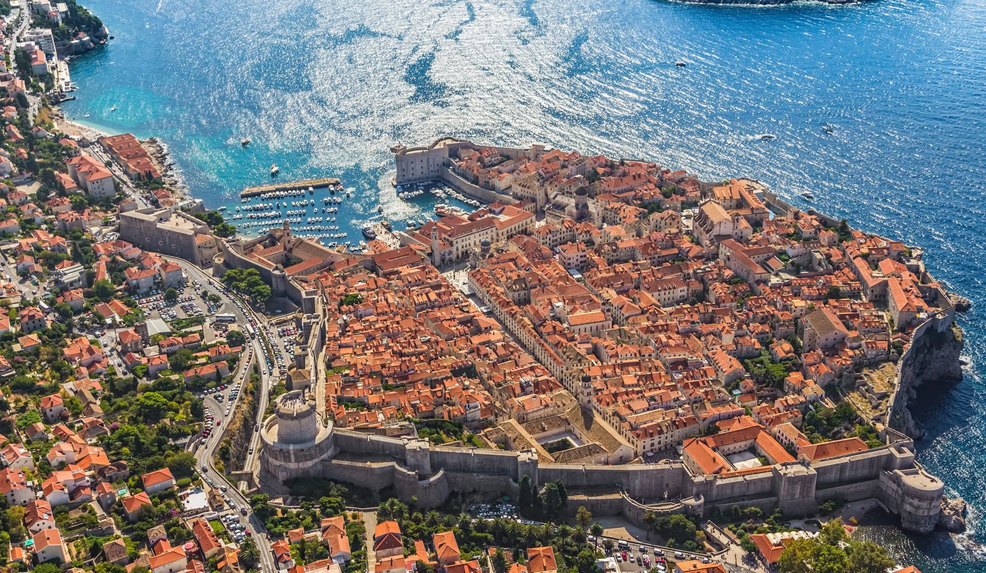 Aerial view of Dubrovnik Old Town walls and terracotta roofs next to sparkling blue Adriatic Sea.
