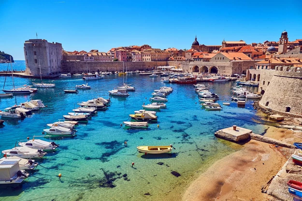 Boats moored in clear turquoise water outside Dubrovnik harbor walls under a bright blue sky.