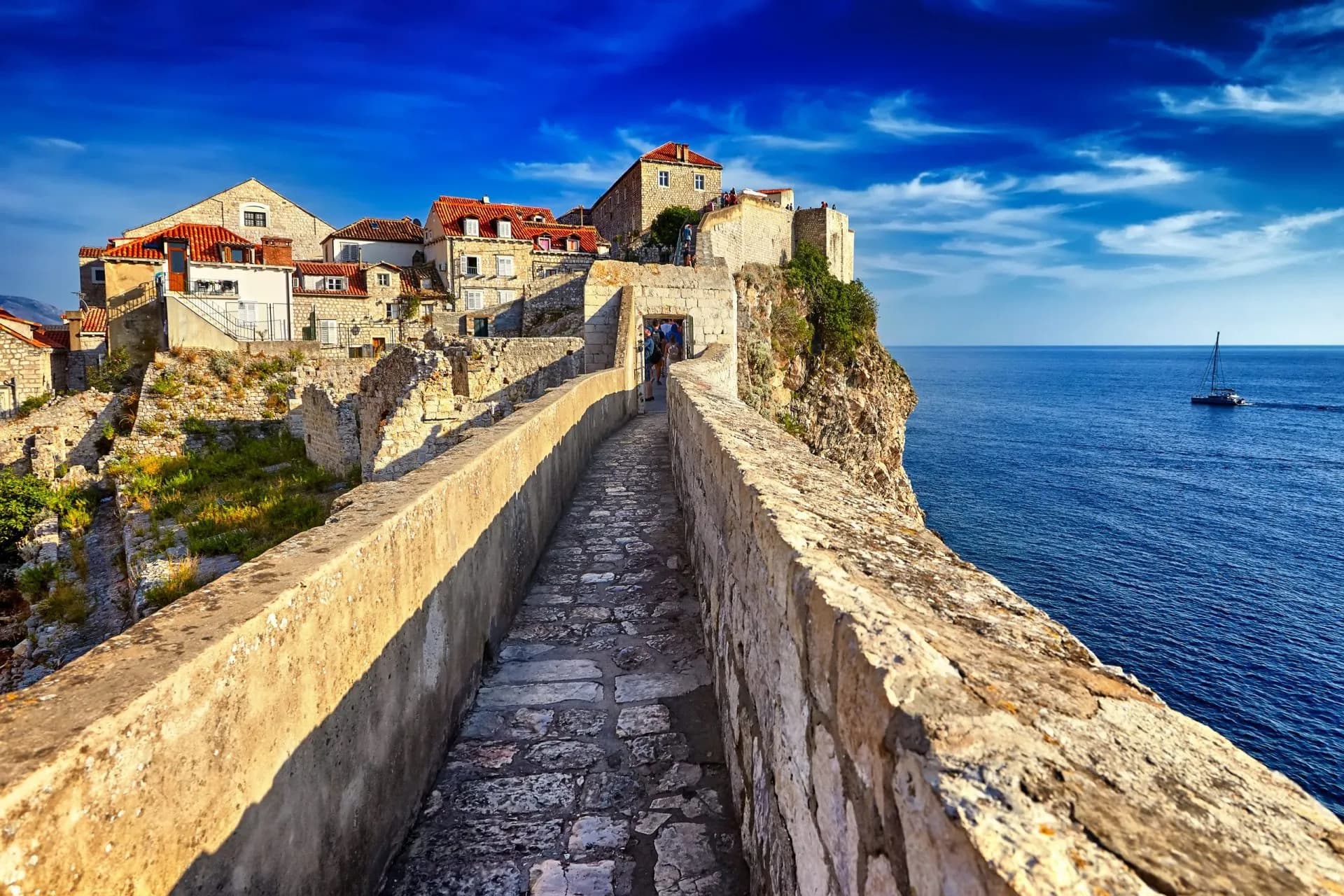 Stone walls of Dubrovnik Old Town overlooking the sea with a sailboat at sunset.