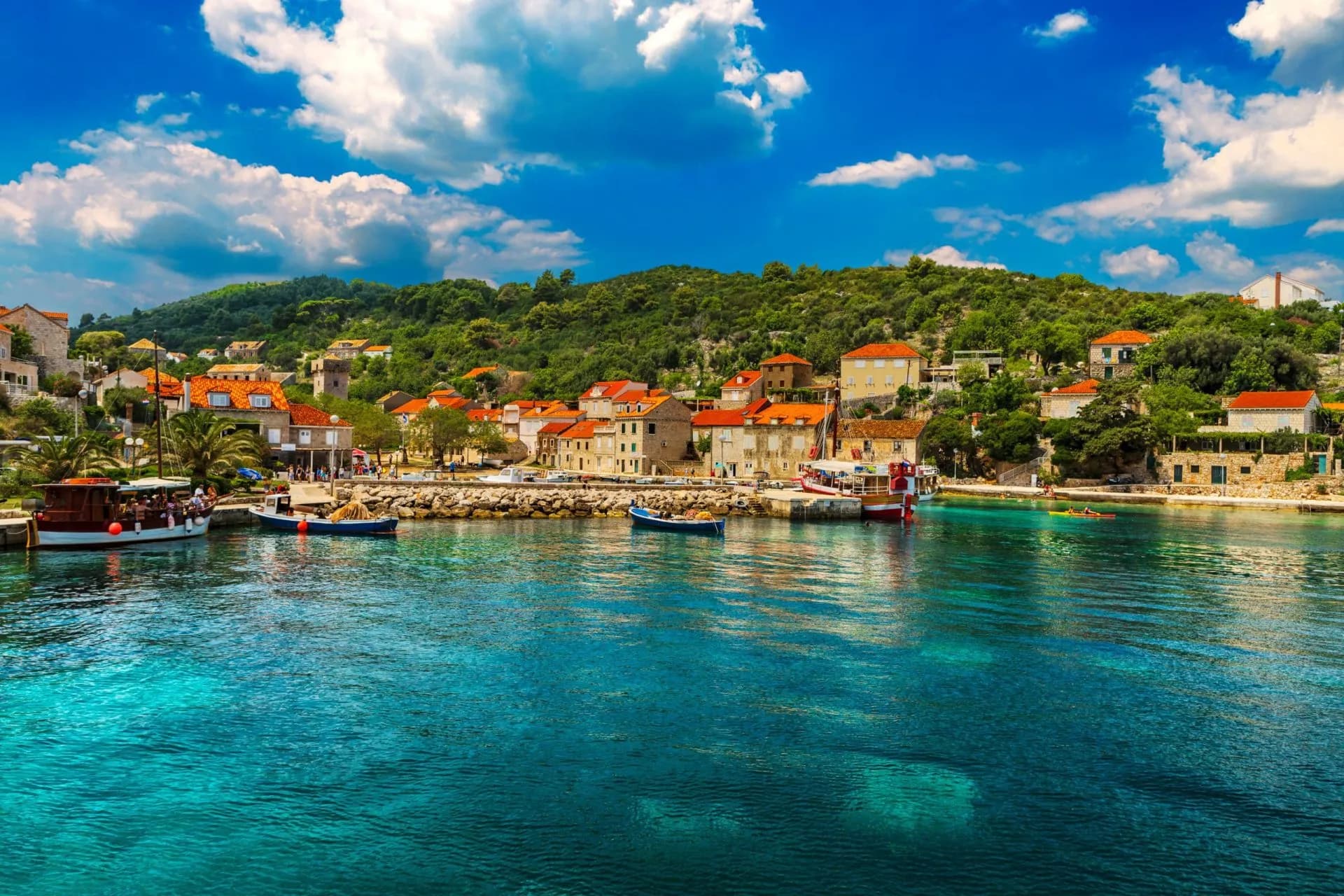 Boats docked in harbor with turquoise water, village on green hillside under blue sky, Sipan Island.