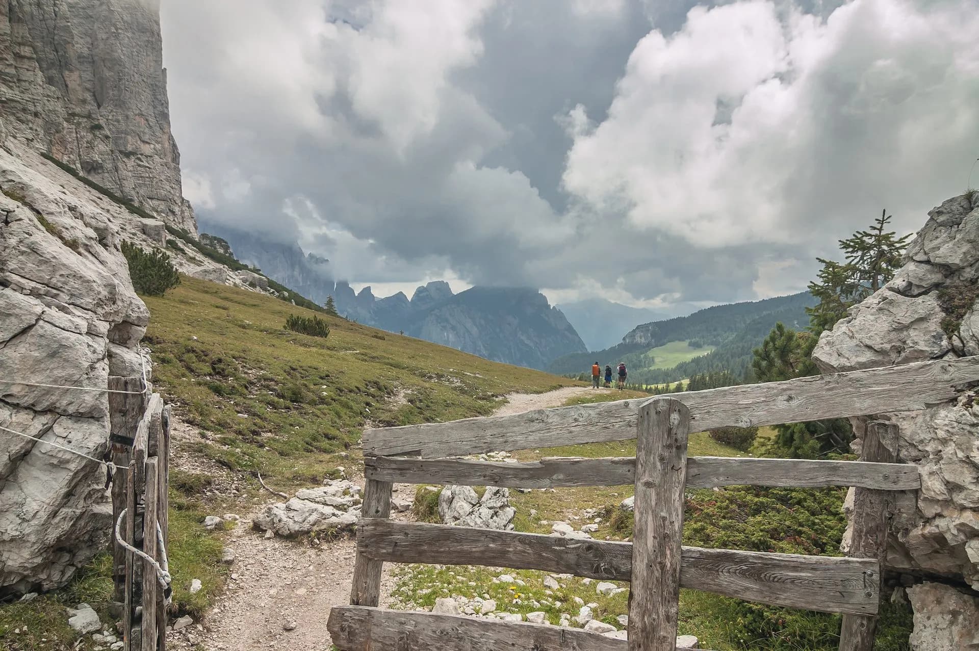 Hikers on trail past wooden fence with dramatic mountain peaks under cloudy sky on Alta Via 1.