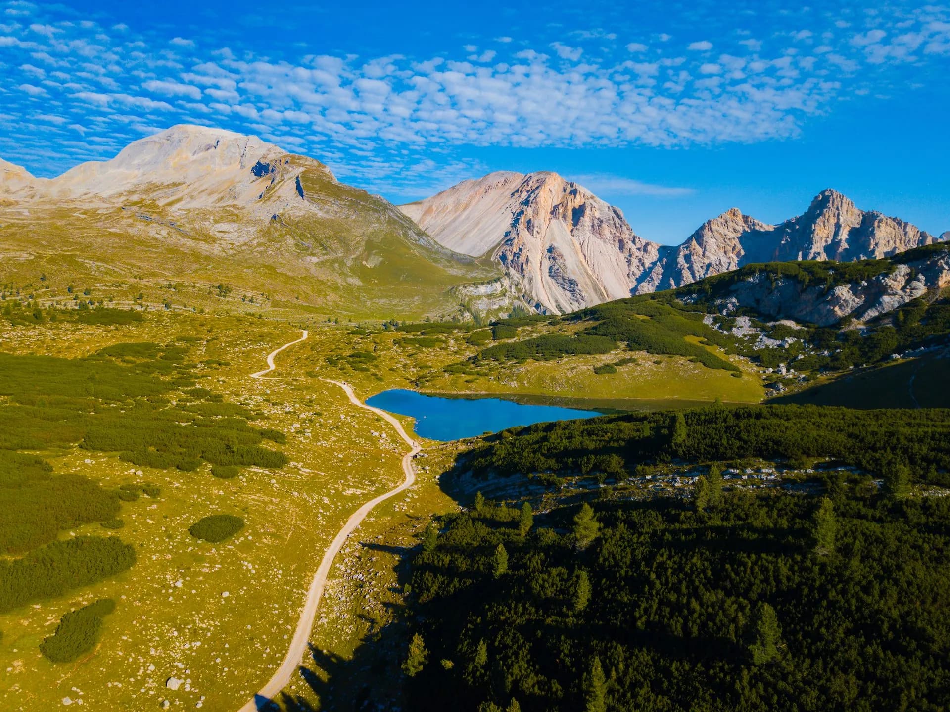 Alpine lake with bright blue water, dirt path, and rugged mountains under a blue sky.
