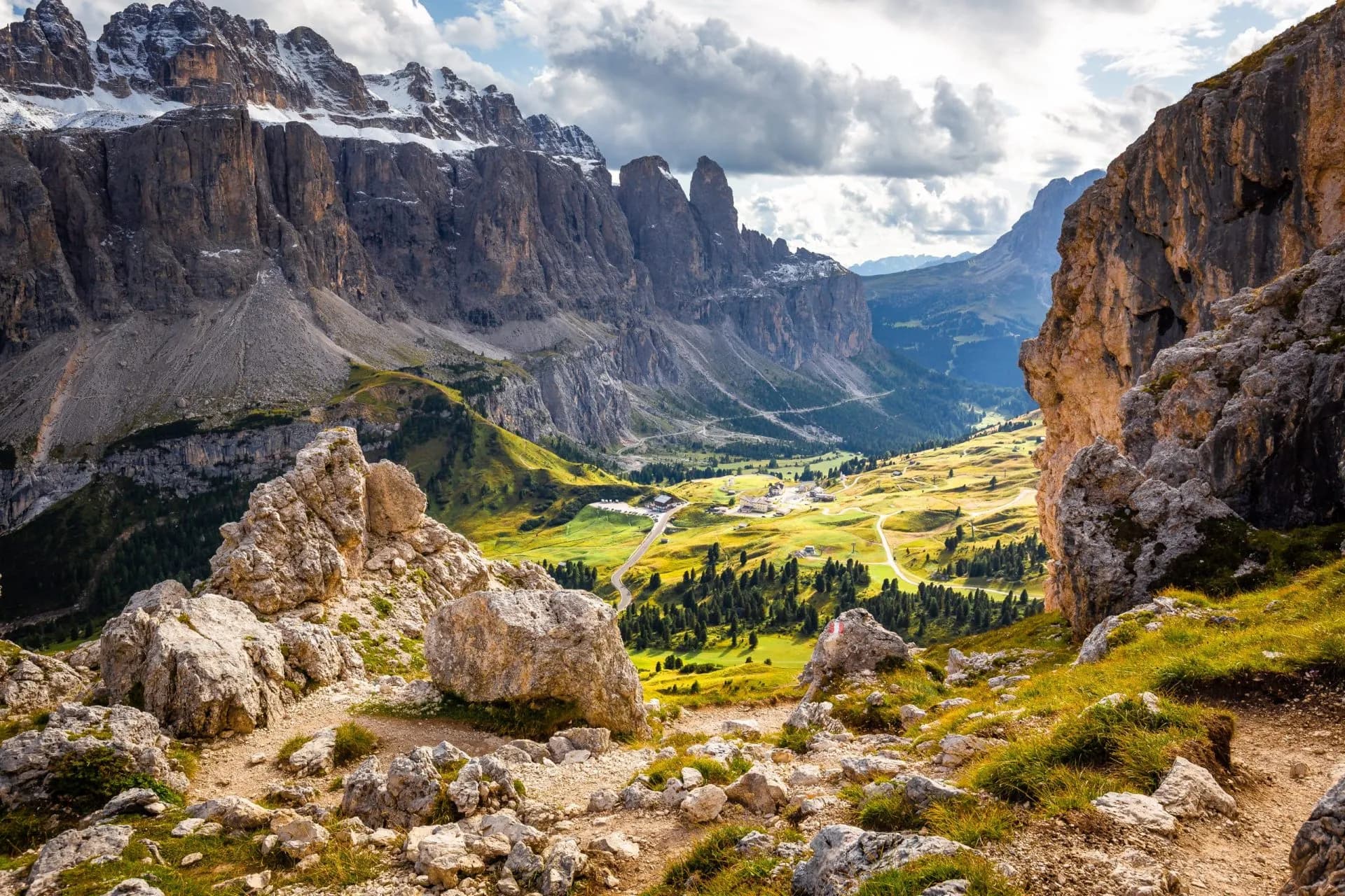Hiking trail overlooking jagged Dolomites peaks with snow, green valley, and village below.
