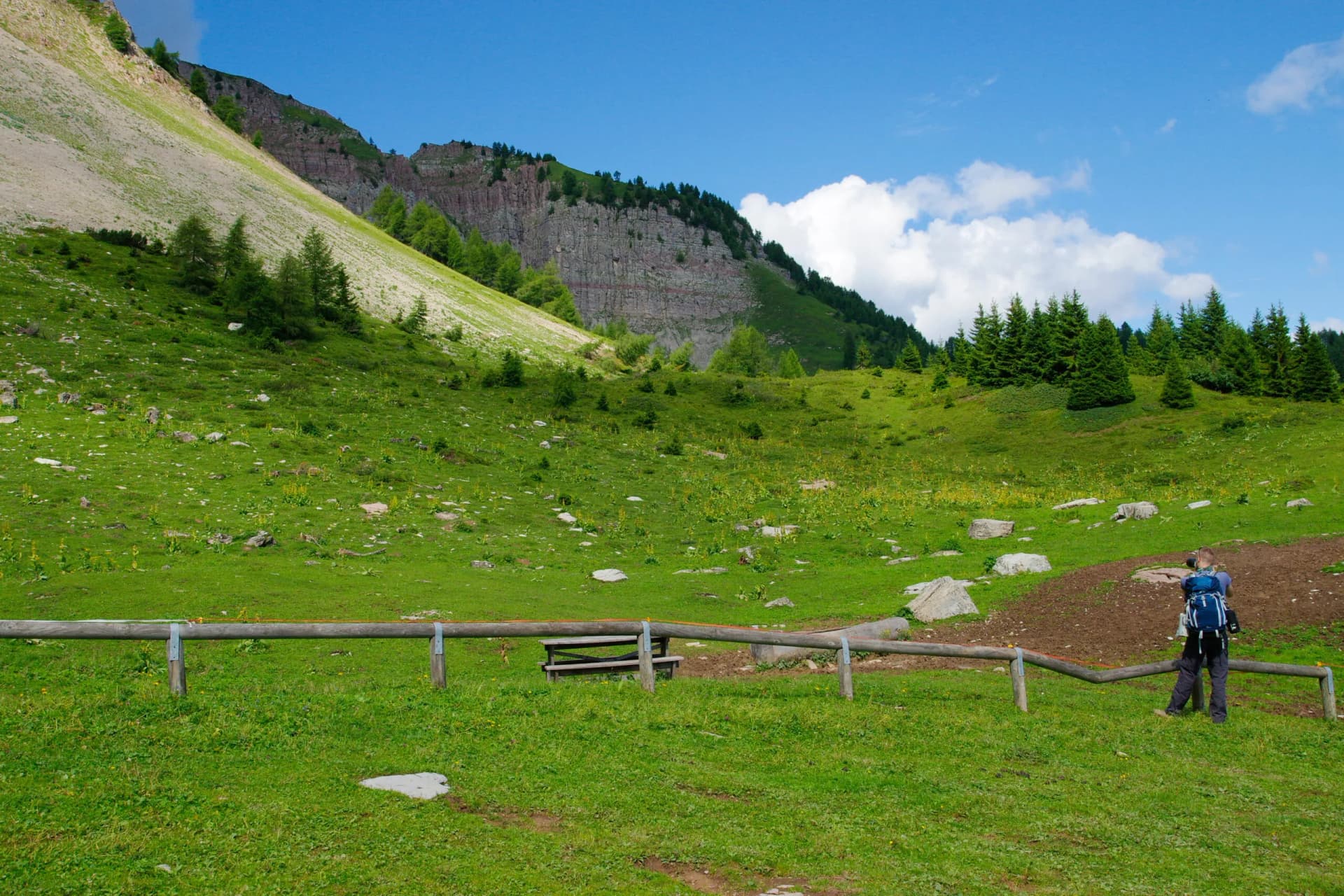 Hiker with backpack photographing green meadows and steep rocky mountains in Brenta Dolomites.