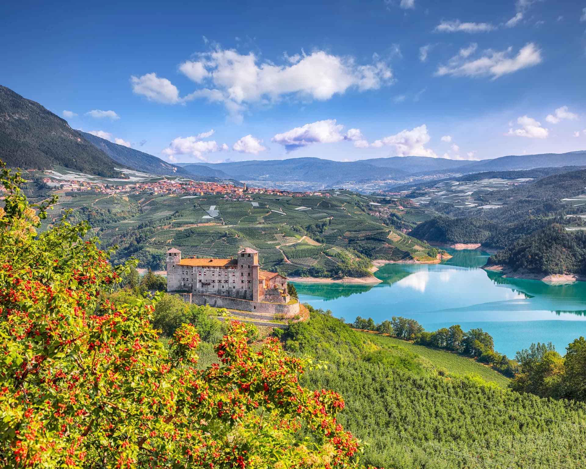 Castle overlooking turquoise lake, terraced orchards, and mountain town under blue sky.