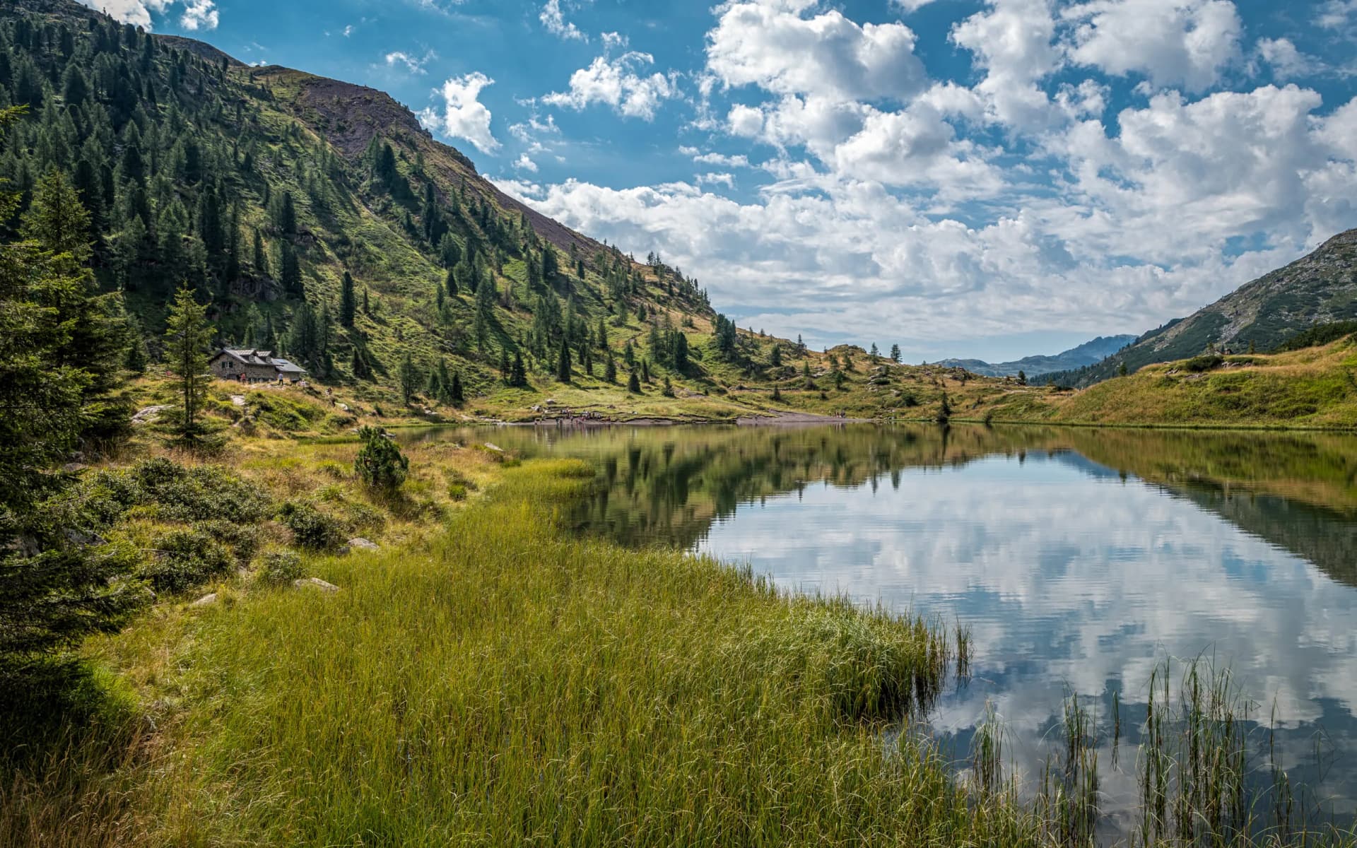 Alpine lake reflecting clouds and mountains, with a stone hut near grassy slopes at Laghi di Colbricon.
