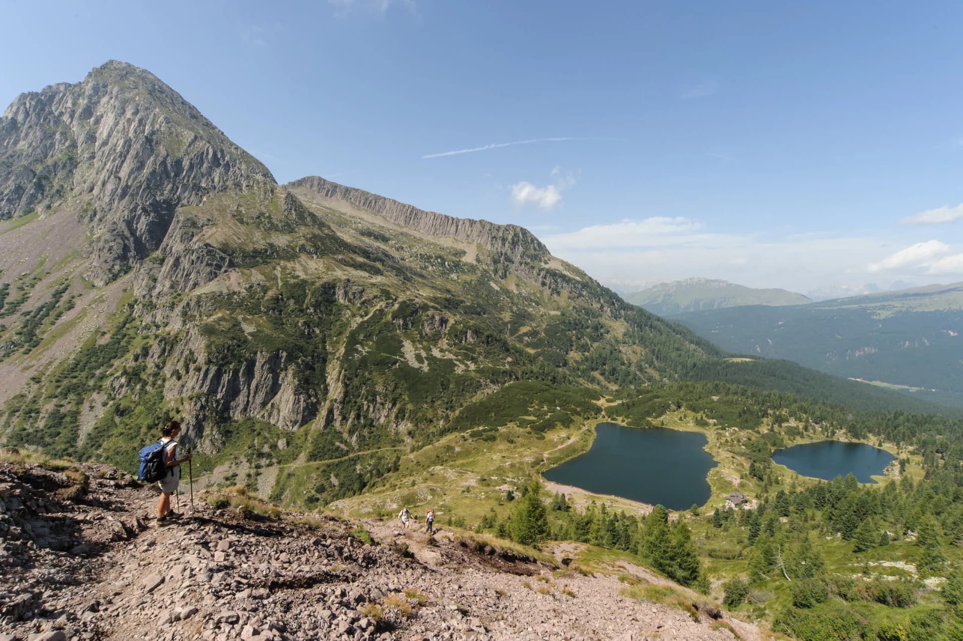 Hikers on rocky trail overlooking Colbricon Lakes and steep mountain slopes under blue sky.