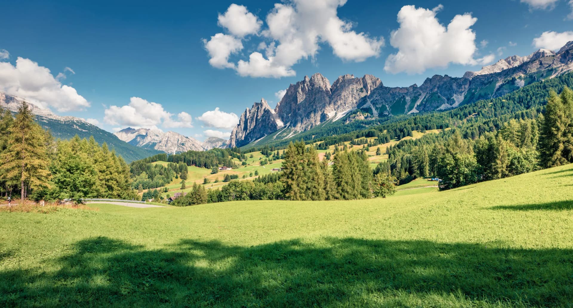 Hiking toward Cortina d'Ampezzo past green meadows and rugged Dolomite mountains under a blue sky.