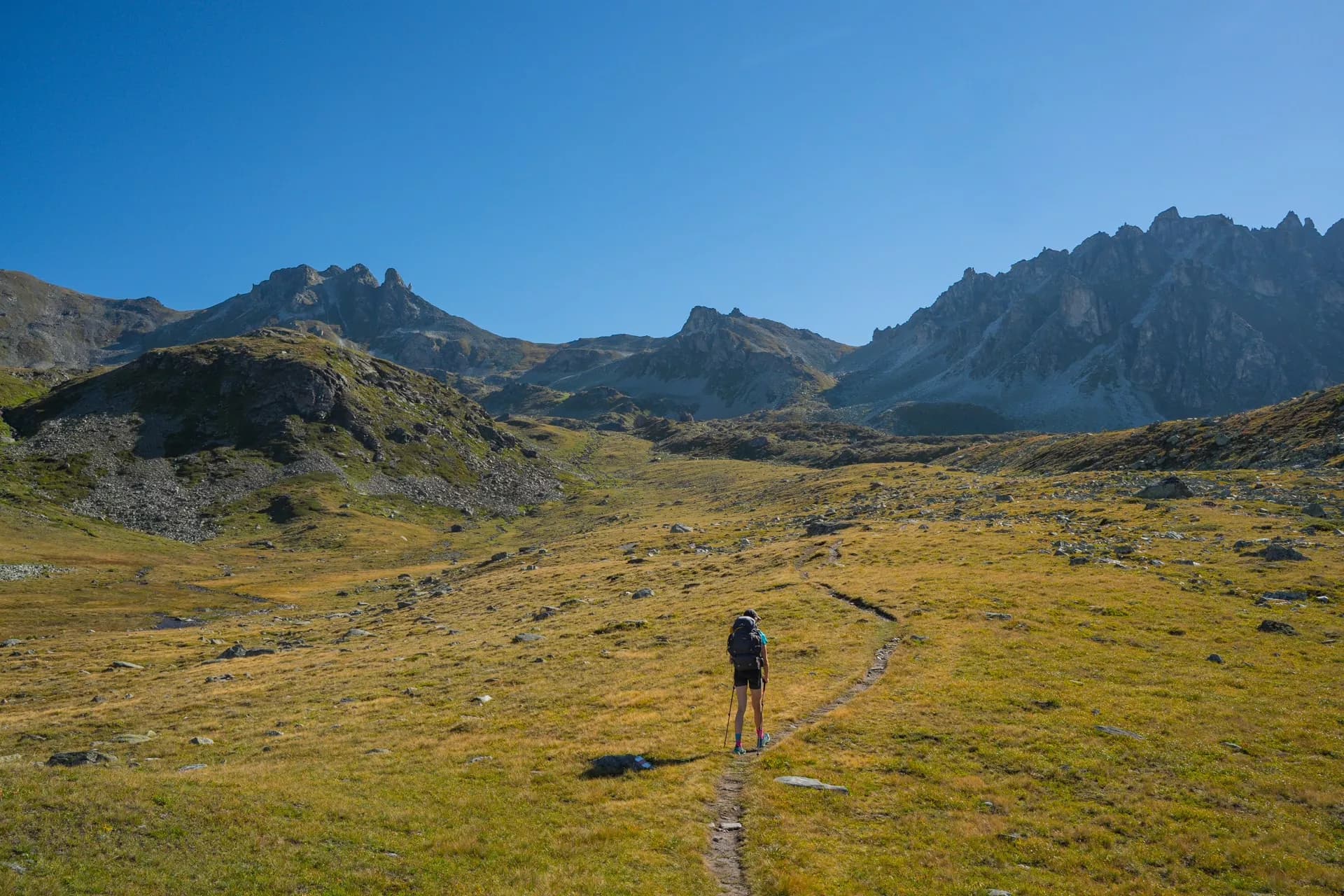 Hiker with trekking poles walking on narrow path through grassy alpine meadow toward rocky mountains.
