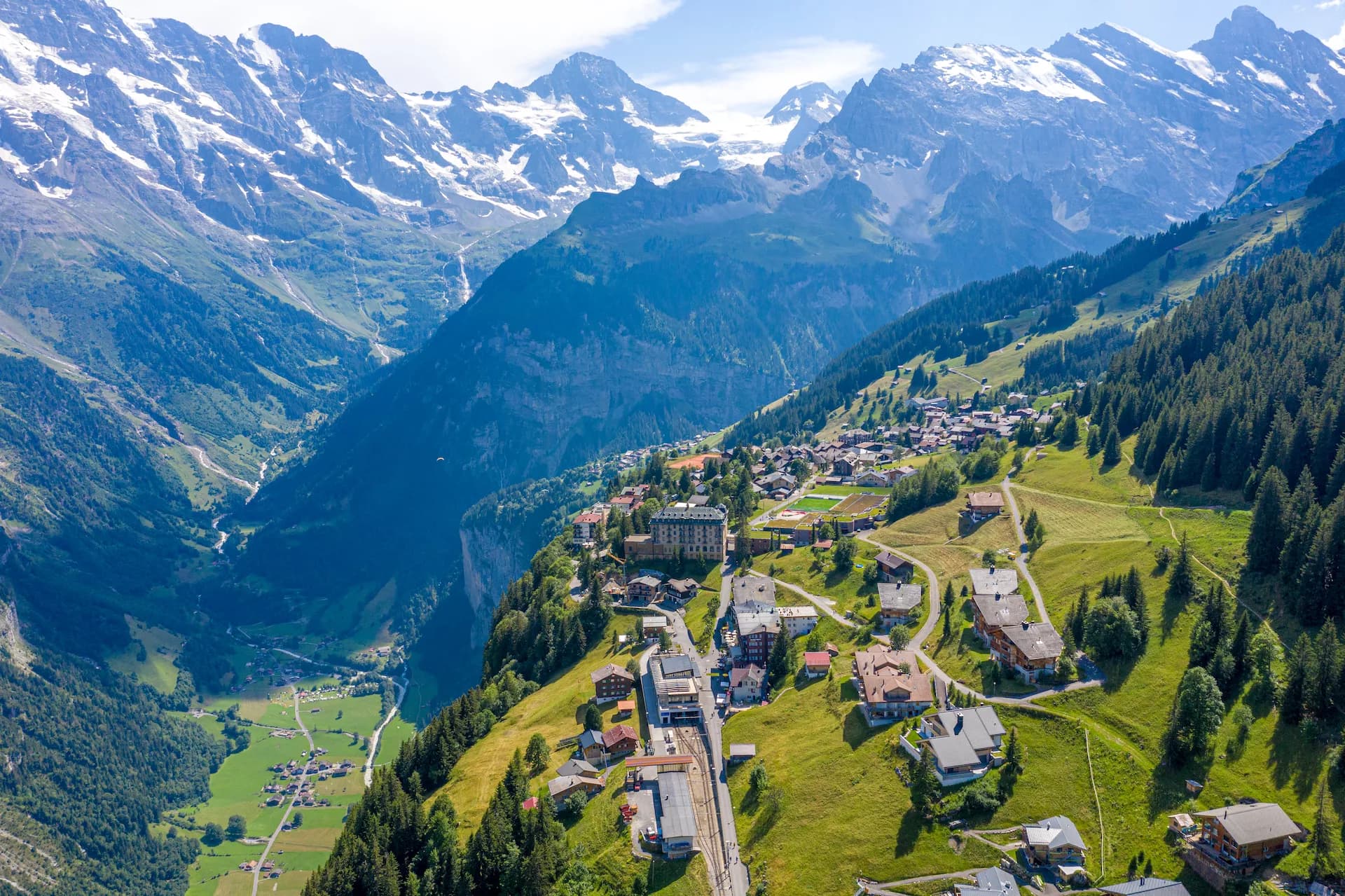 Aerial view of Mürren village nestled on a green slope below snow-capped Swiss Alps.