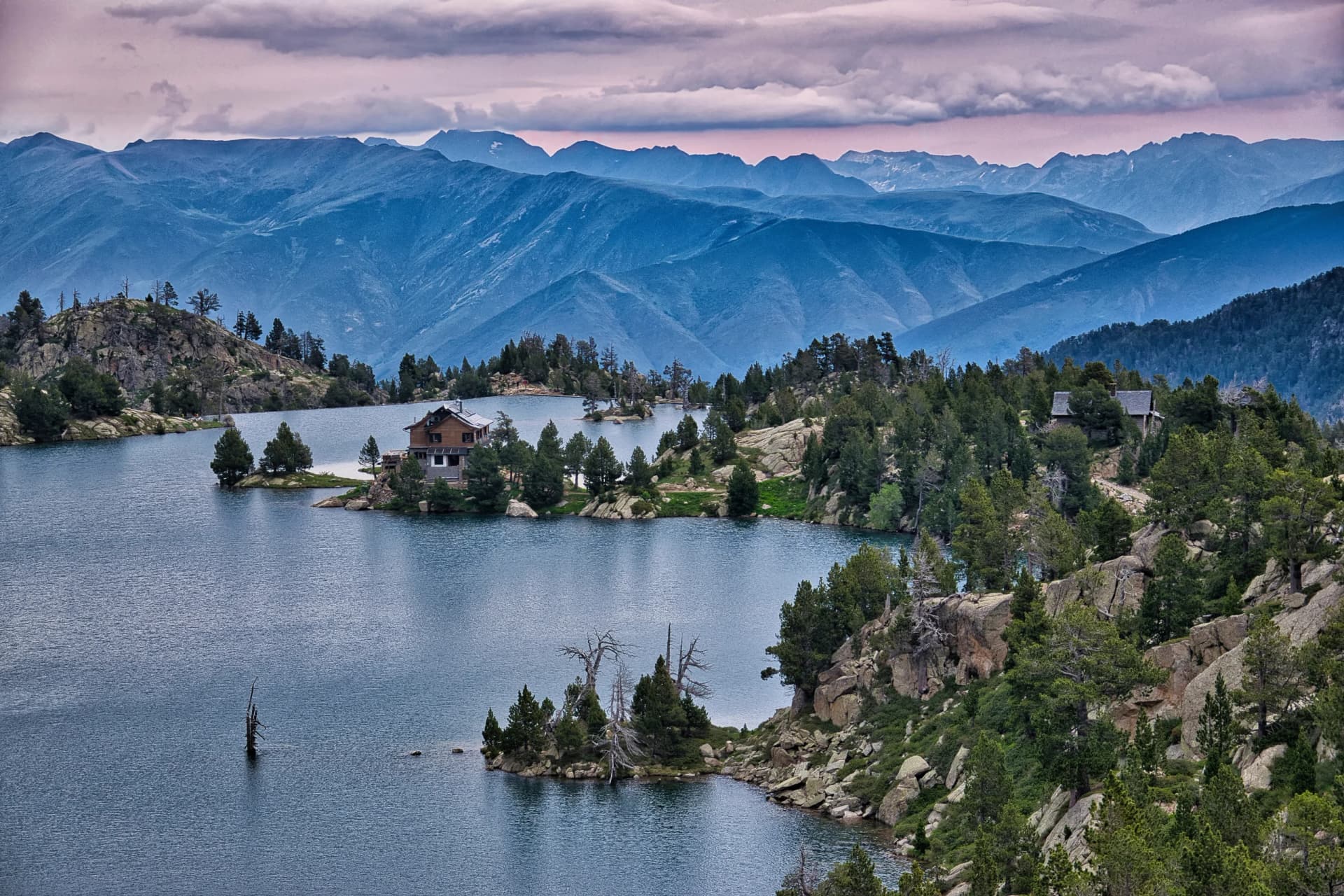 Mountain lake with wooden refuge, rocky islands, and blue mountain range backdrop.