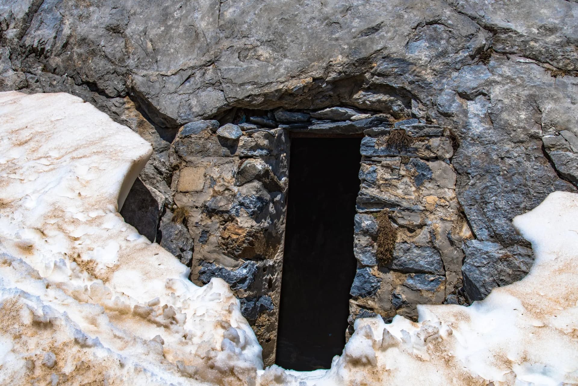 Stone entrance built into a rocky cliff face next to melting, dirty snow.