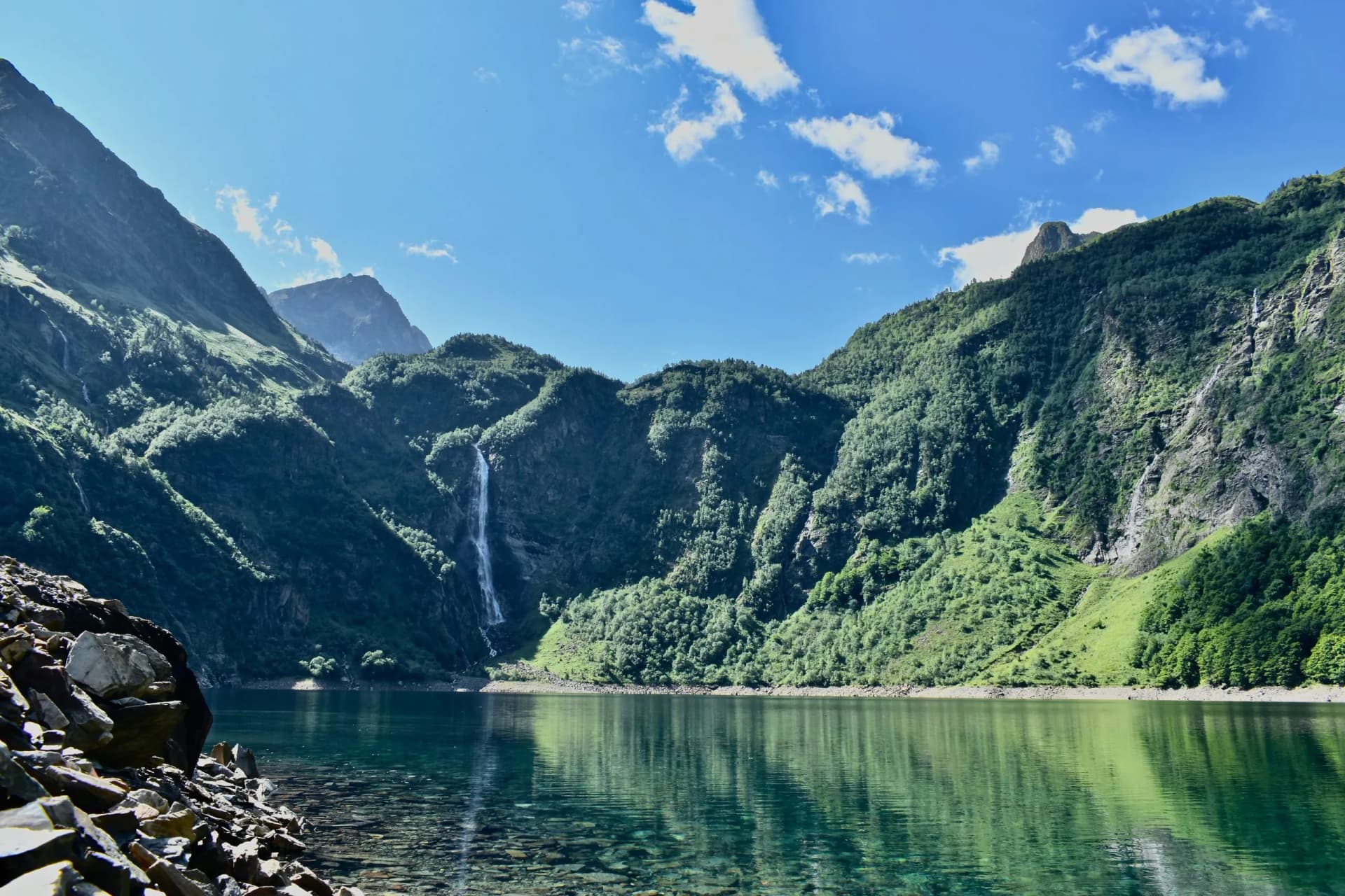 Waterfall cascading down lush green mountainsides into a clear alpine lake under a blue sky.