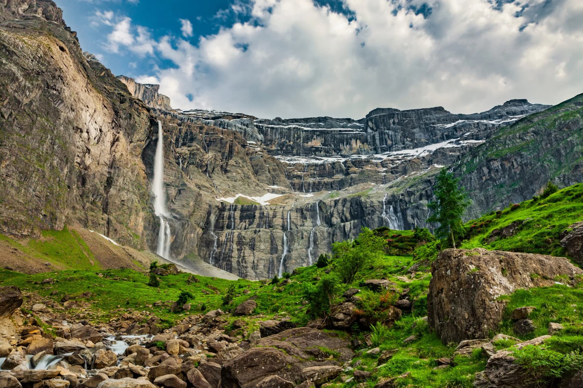 Tall waterfall cascading down massive rock face in Cirque de Gavarnie with green slopes and snow patches.