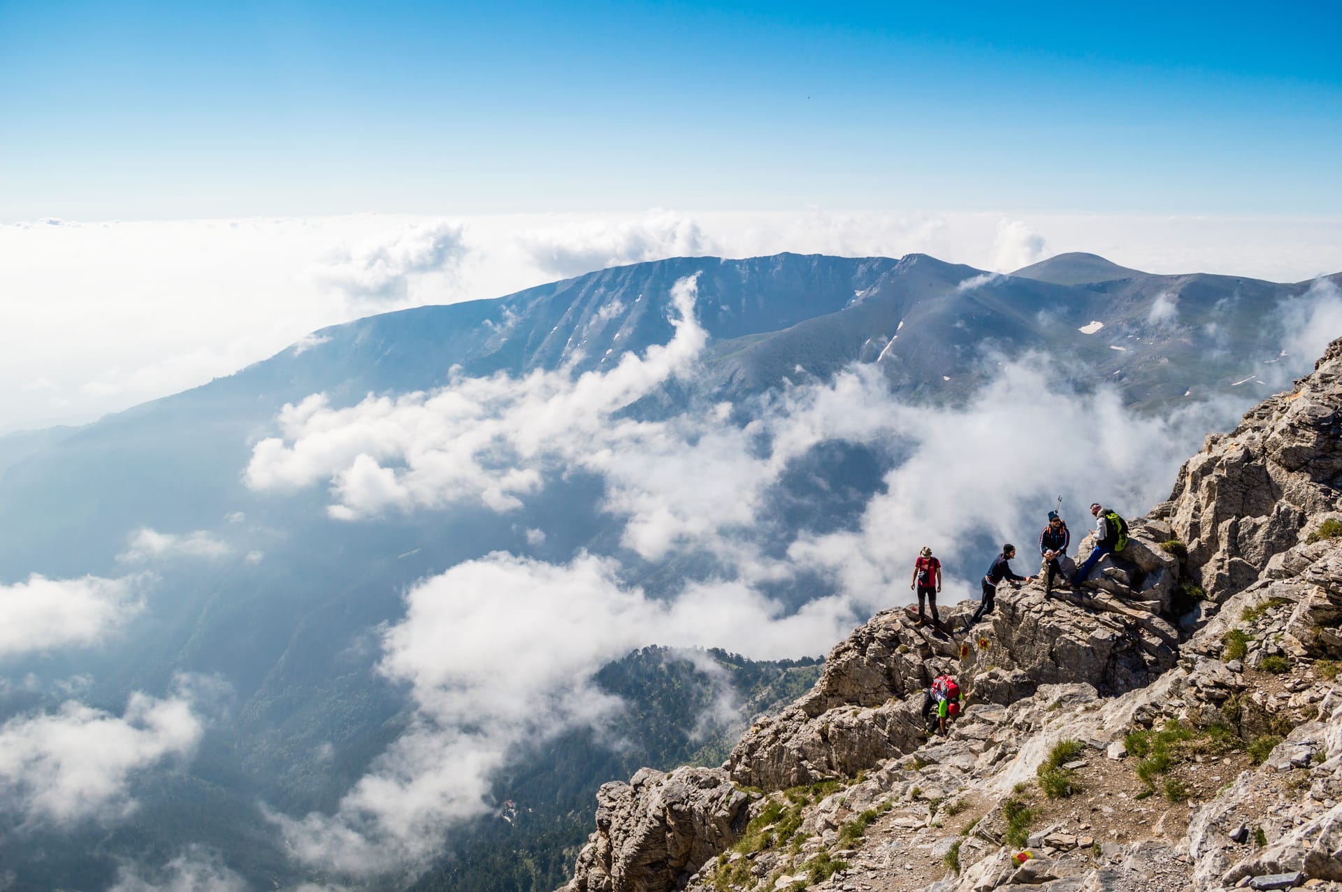 Hikers resting on rocky mountain ridge above clouds with distant peaks under bright blue sky