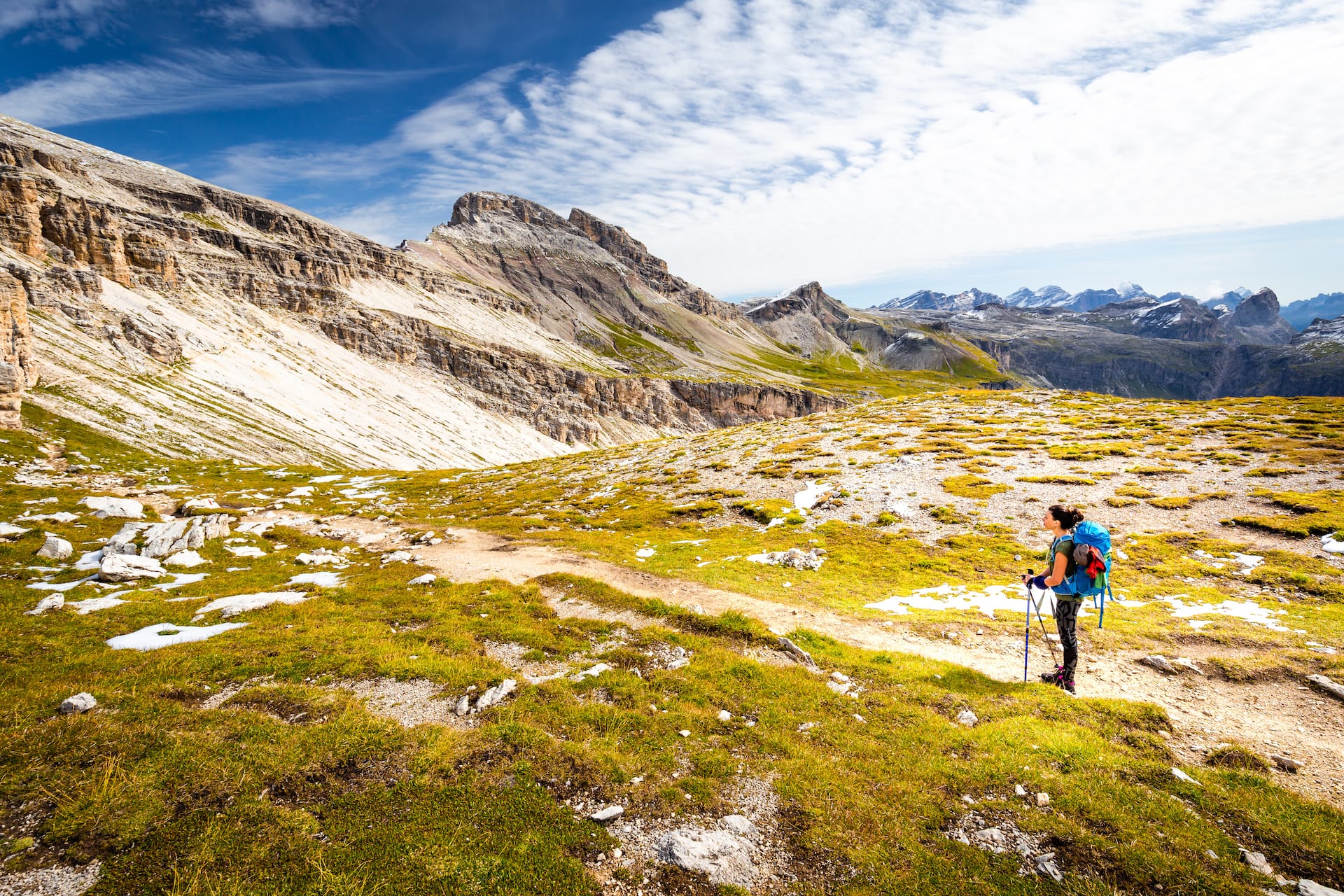 Hiker with backpack and poles pauses on trail amidst alpine meadow with rocky mountains backdrop.