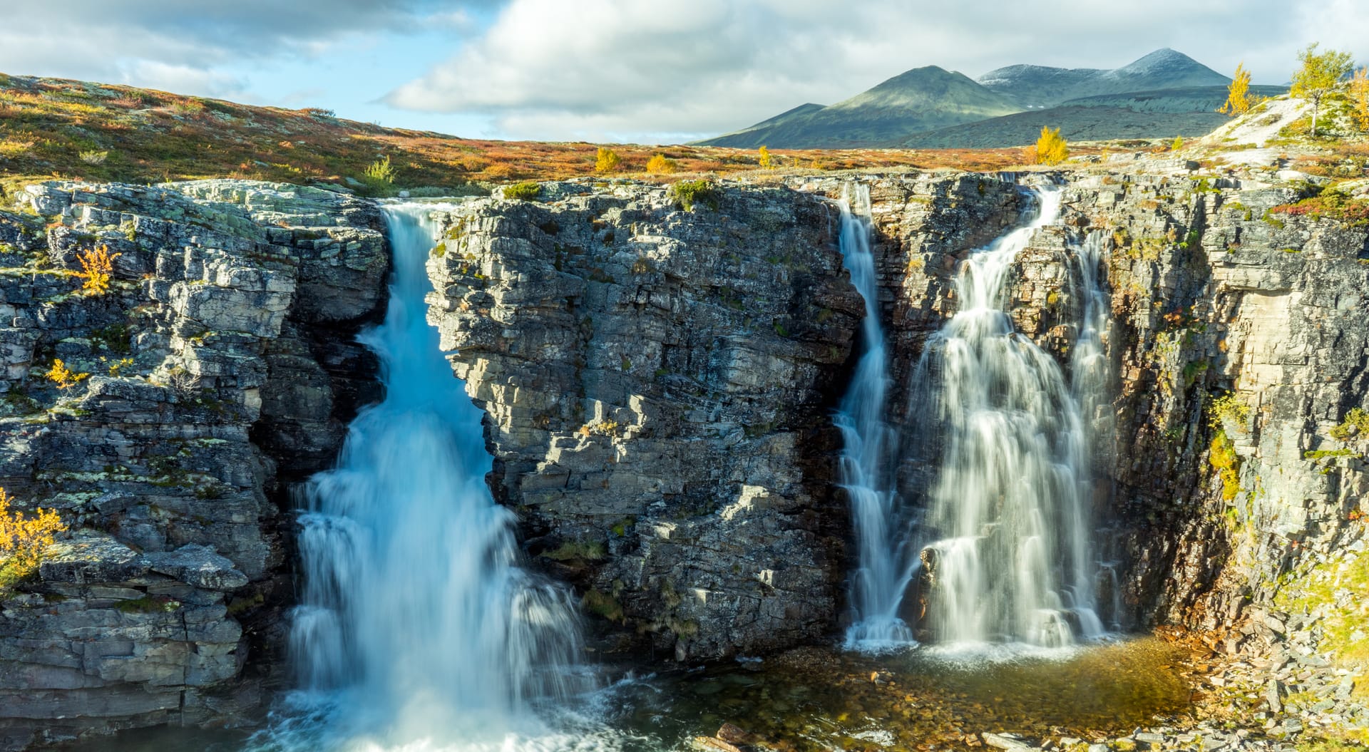 Waterfall cascading over rocky cliffs with autumn foliage and mountains in the background, Norway.